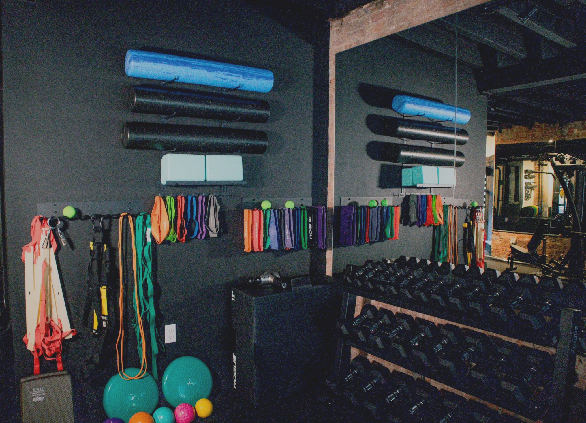 Exercise equipment in a gym, including kettlebells, resistance bands, foam rollers, medicine balls, and a rack of dumbbells, with black walls and some mirrors in the background.