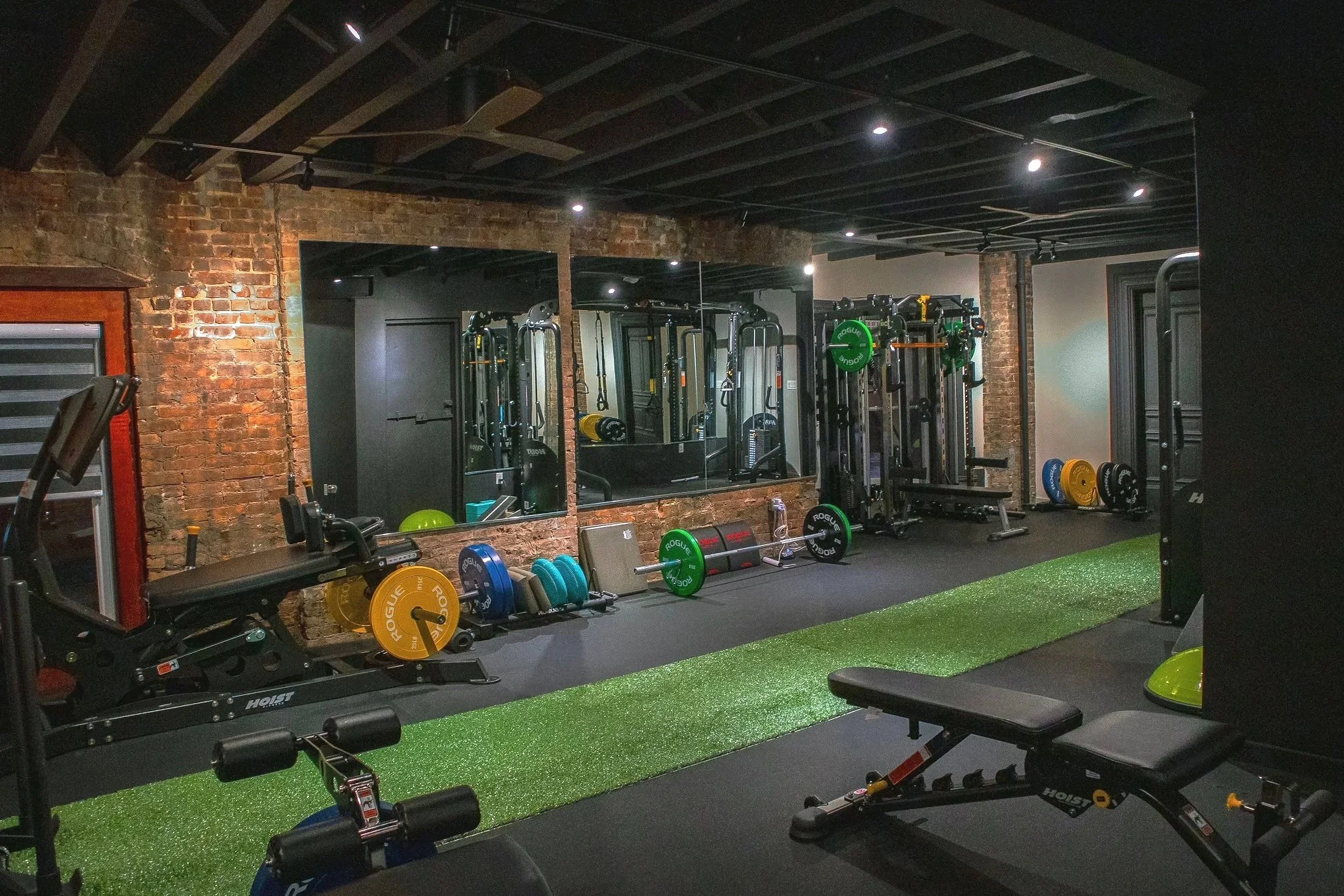 Indoor gym with weightlifting equipment, including barbells, weight plates, and benches, brick walls, mirrors, and a green artificial turf strip.