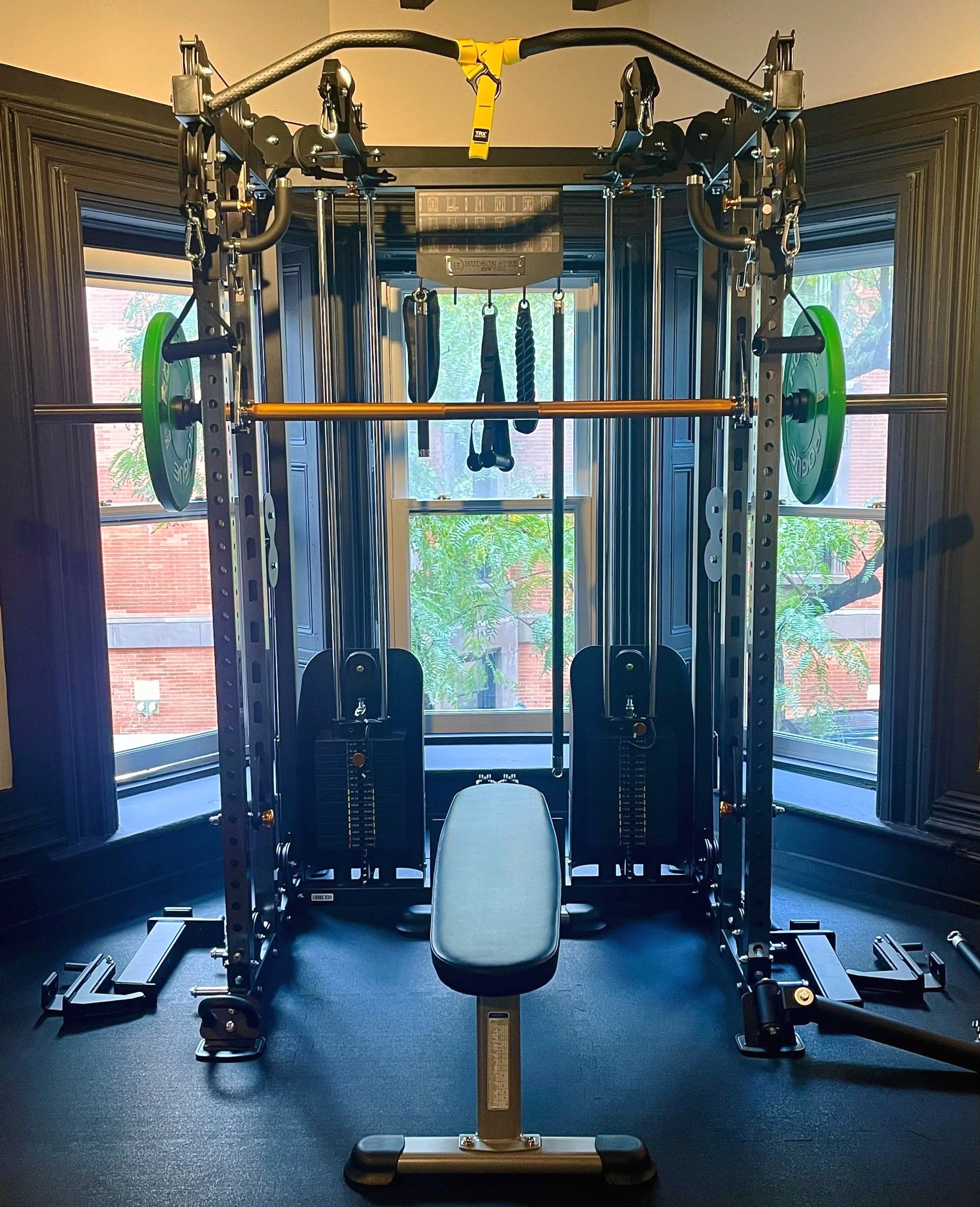 A home gym setup with a power rack, barbell, weight plates, weight stack, adjustable bench, and workout straps in front of two windows with trees outside.