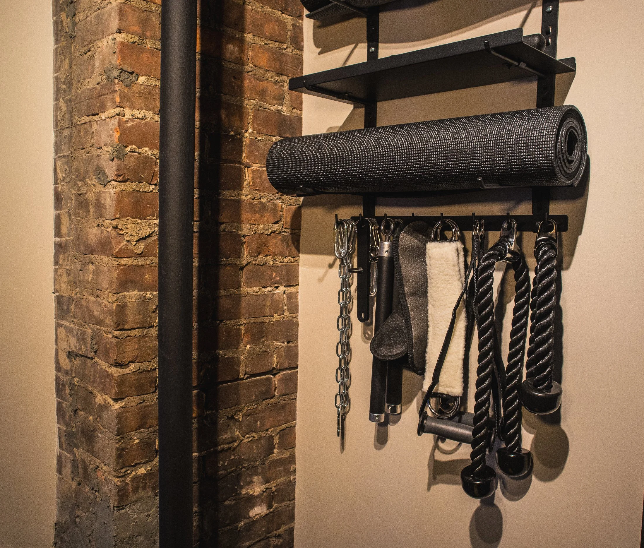 Wall-mounted organizer with workout accessories including a yoga mat, ropes, chains, and other exercise tools in a room with exposed brick wall.