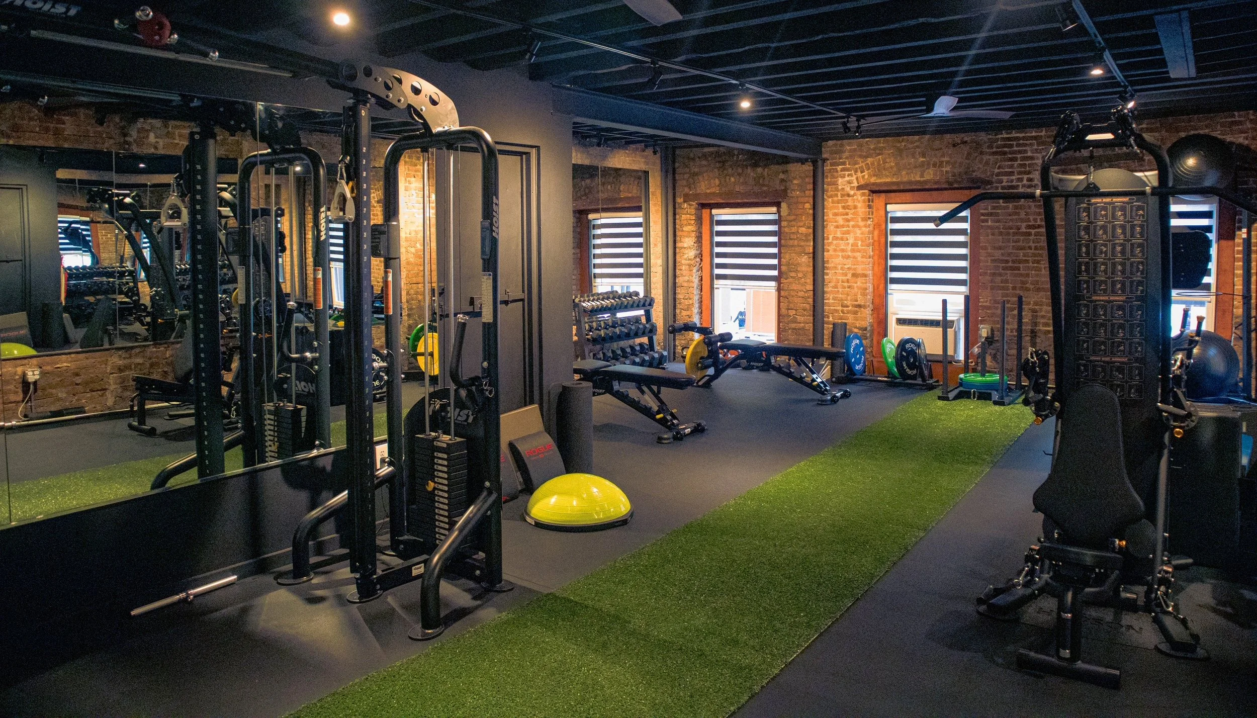 Interior view of a gym with various exercise equipment, including weights, a balance ball, and a plyometric box, with brick walls and windows with horizontal blinds.
