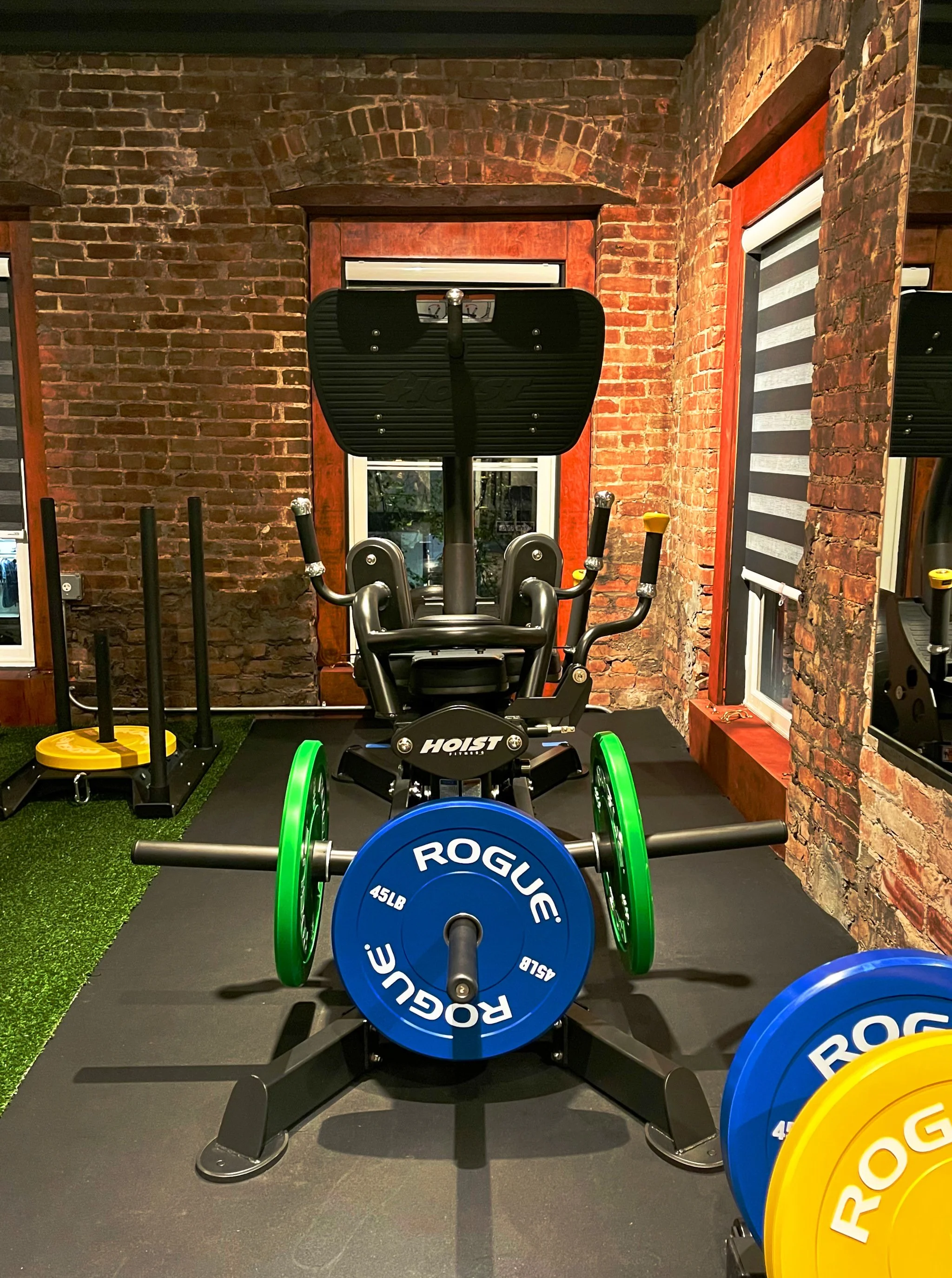 Gym with weightlifting equipment, including a barbell with blue and yellow weight plates labeled 'ROGUE' and '45LB', a black incline workout machine, and a small sled with black handles on a rubber mat, brick walls, windows, and a mirror.