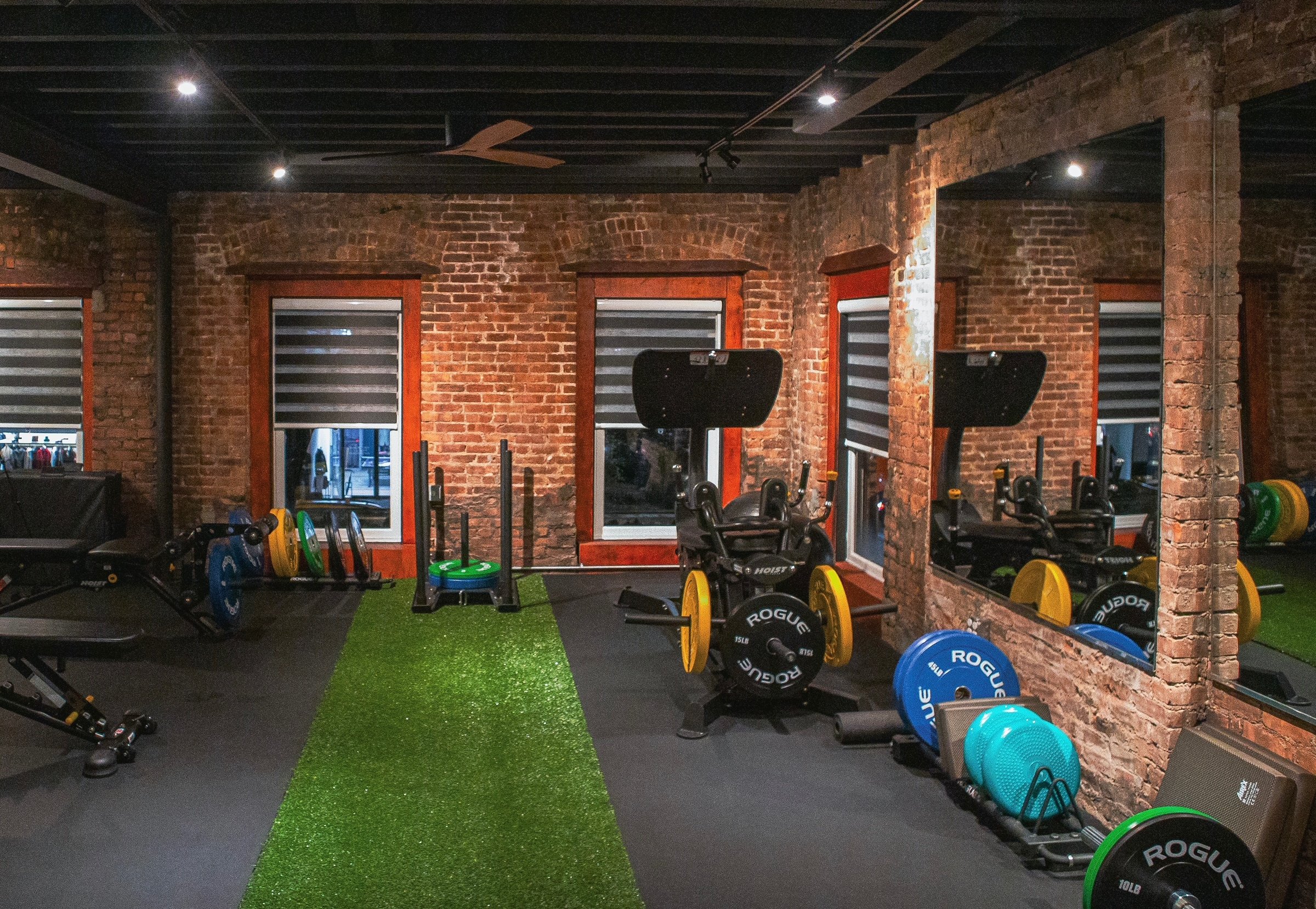 Indoor gym with brick walls, three windows with wooden frames, black ceiling, dim lighting, artificial turf in the center, and various weightlifting equipment including barbells, weights, and a leg press machine.