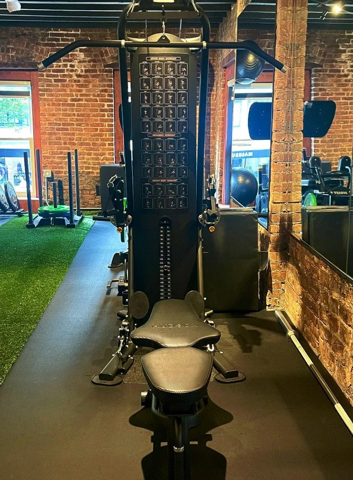 Interior of a gym with a multi-functional workout machine on a black mat, surrounded by brick walls and some exercise equipment, with windows letting in natural light.