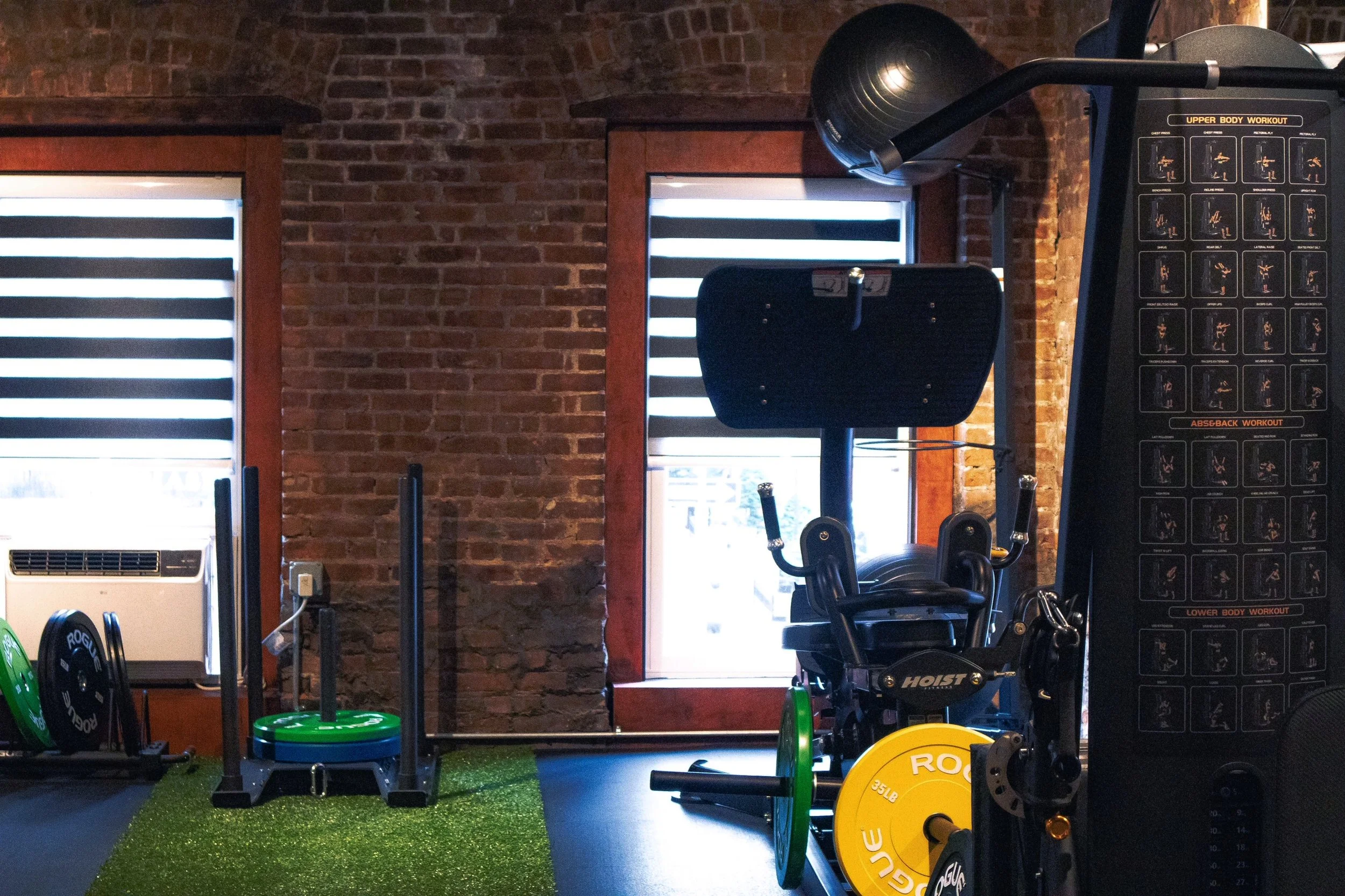 Indoor gym with brick walls featuring workout equipment including a weight machine, weight plates, and a green turf area near two windows with blinds.