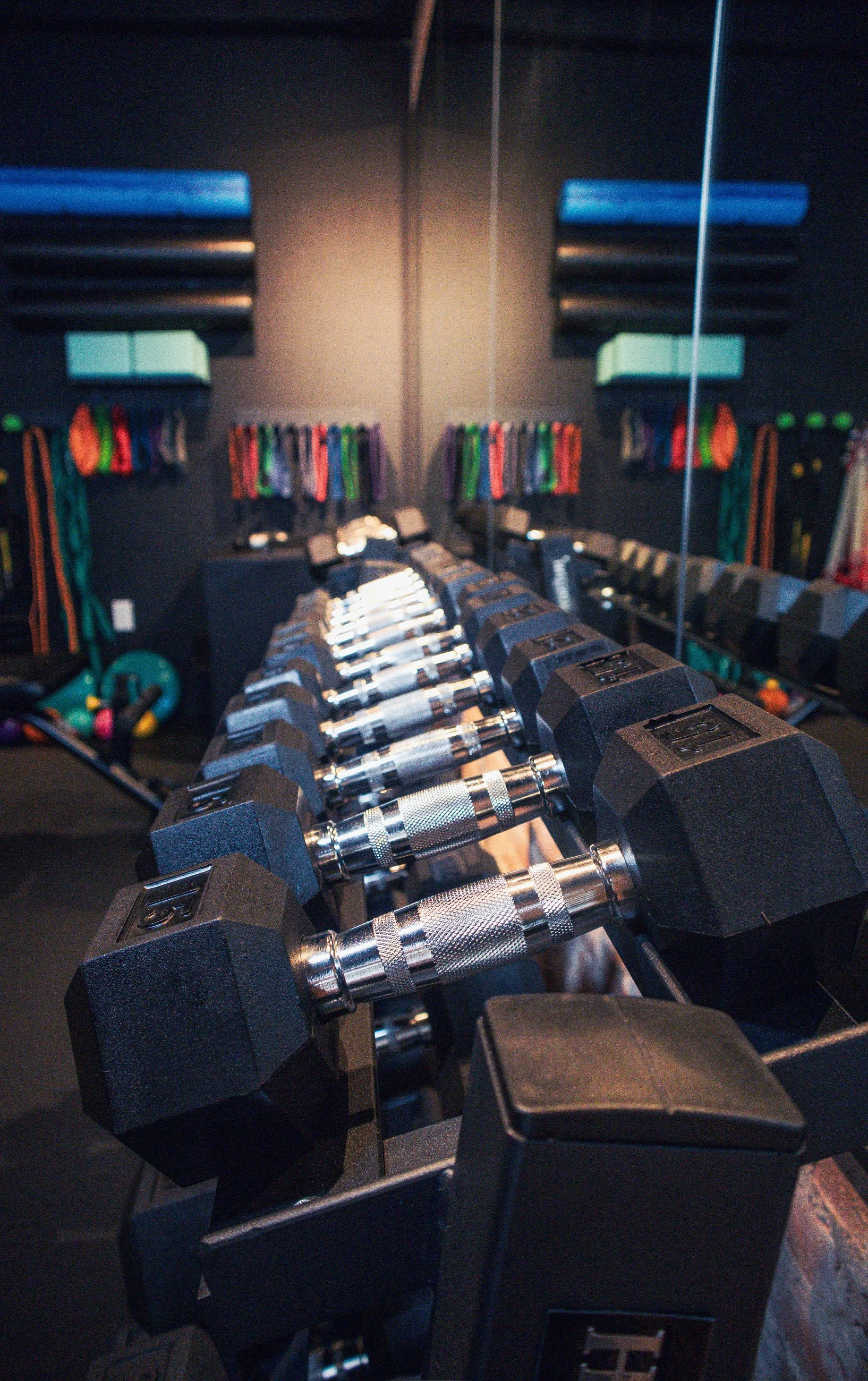 A set of black dumbbells organized on a rack in a gym with a mirror reflecting colorful resistance bands hanging on the wall behind.