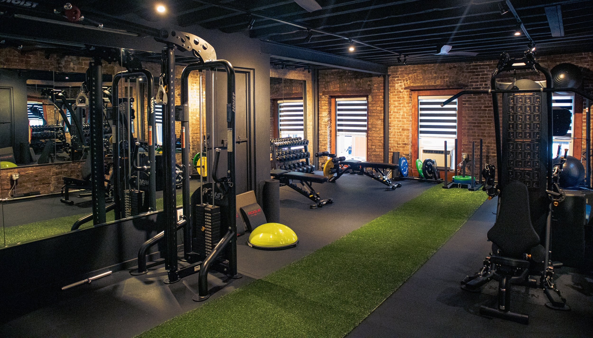 Interior of a gym with various workout equipment including weights, benches, and machines, brick walls, and windows with blinds.