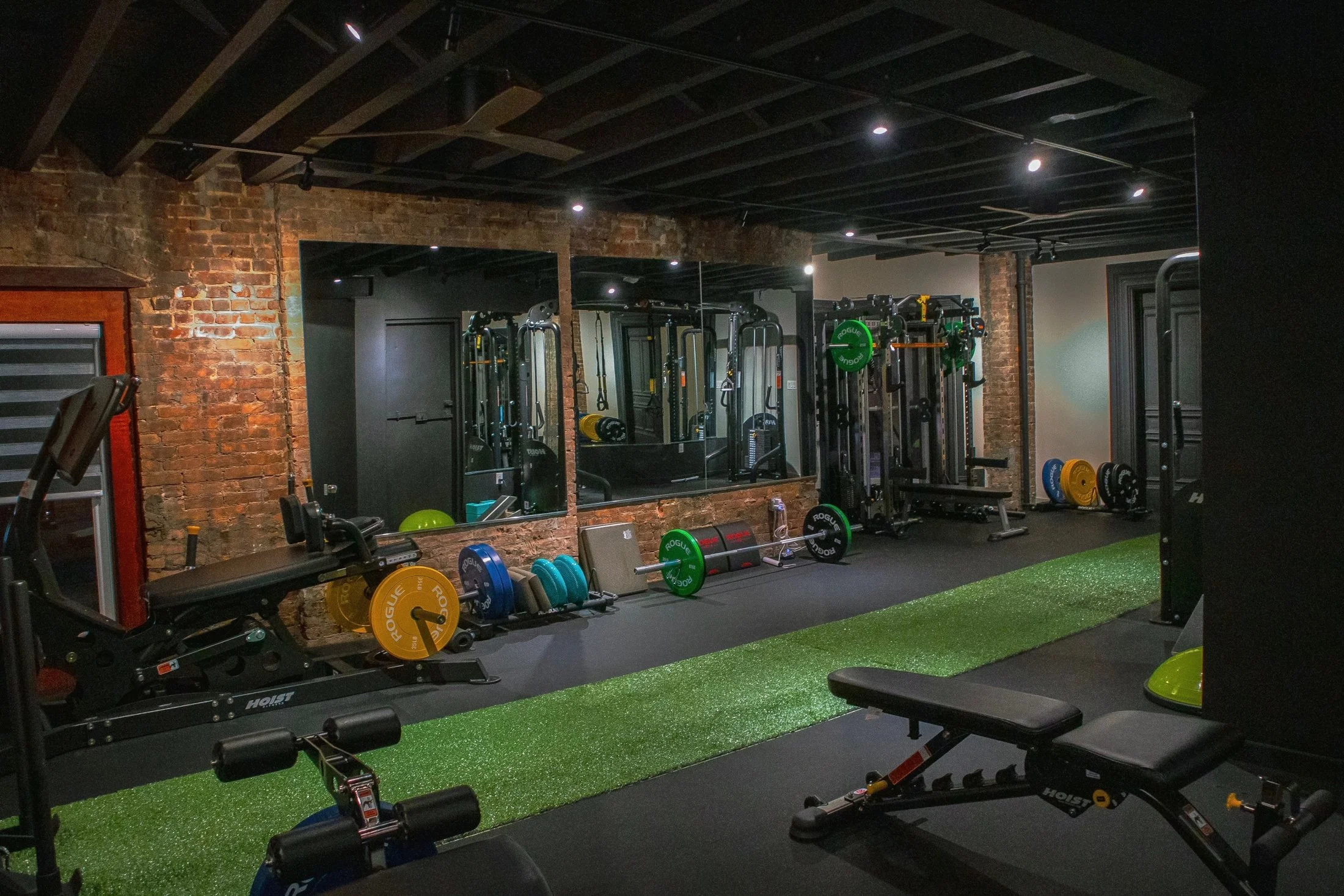 Empty gym with brick walls and black ceiling, equipped with weight machines, barbells, dumbbells, a weight bench, and a green artificial turf section.