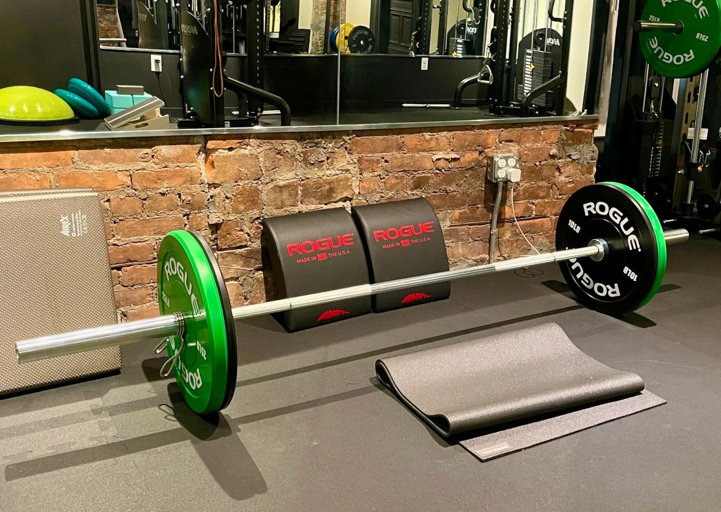 A barbell with green weight plates resting on a gym floor, with a black mat nearby, in front of a brick wall with gym equipment and a mirror.