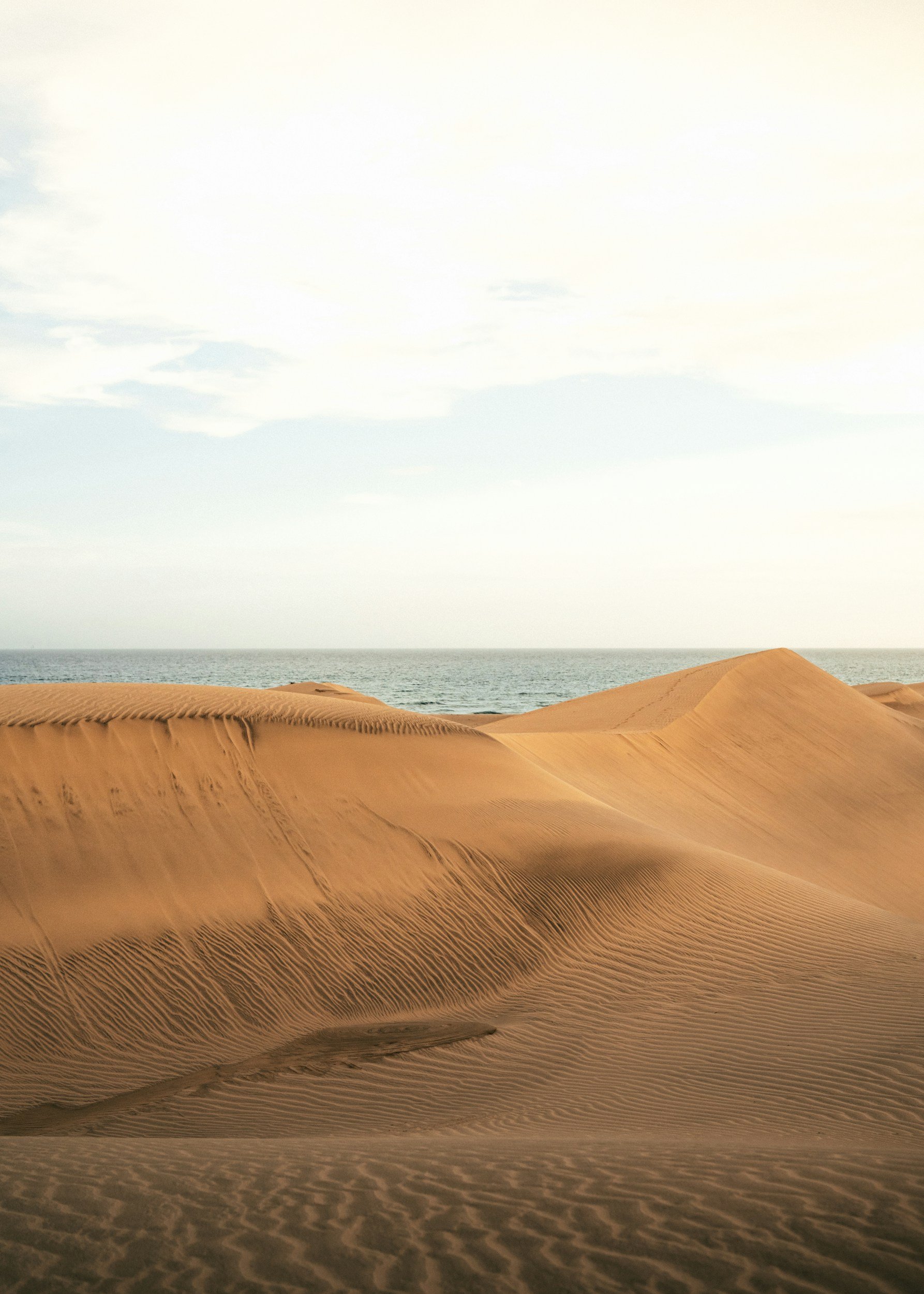 Sand dunes with ripples and patterns near the ocean under a partly cloudy sky.