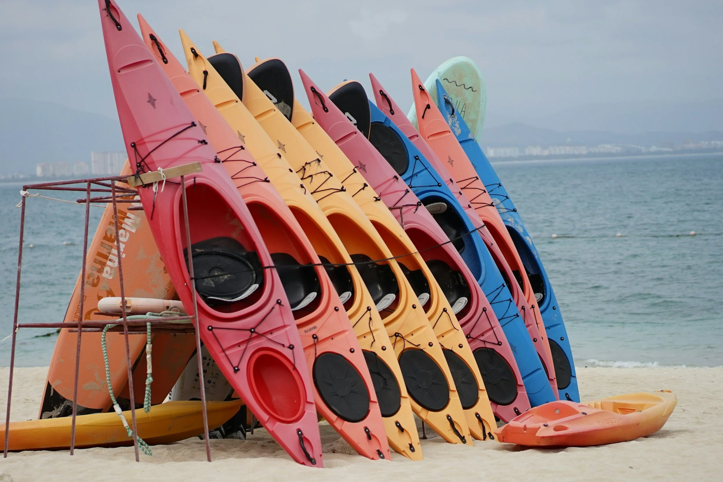 Colorful kayaks stacked on a beach near the water, with a paddleboard lying on the sand nearby.