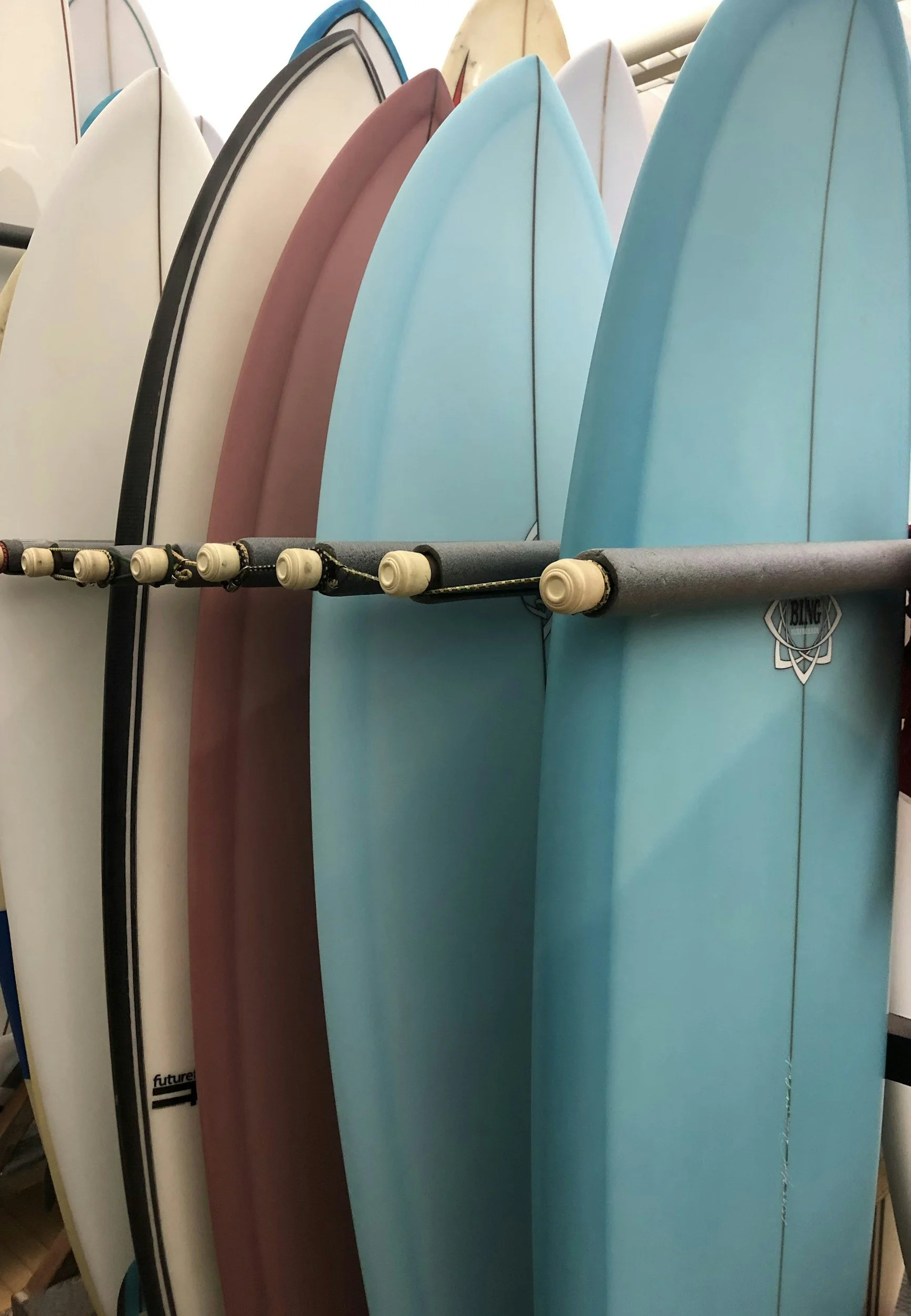 Multiple surfboards of various colors, including white, black, pink, and blue, are standing upright on a rack in a store.