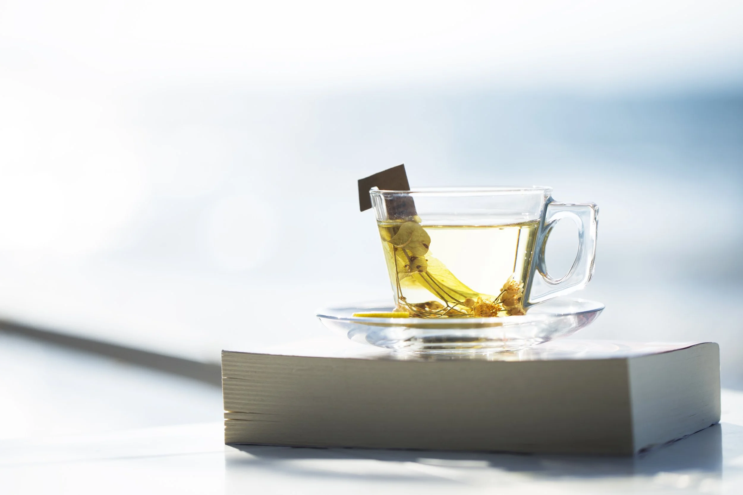 A clear glass cup filled with herbal tea on a glass saucer, placed on a closed book, with a blurred bright background.