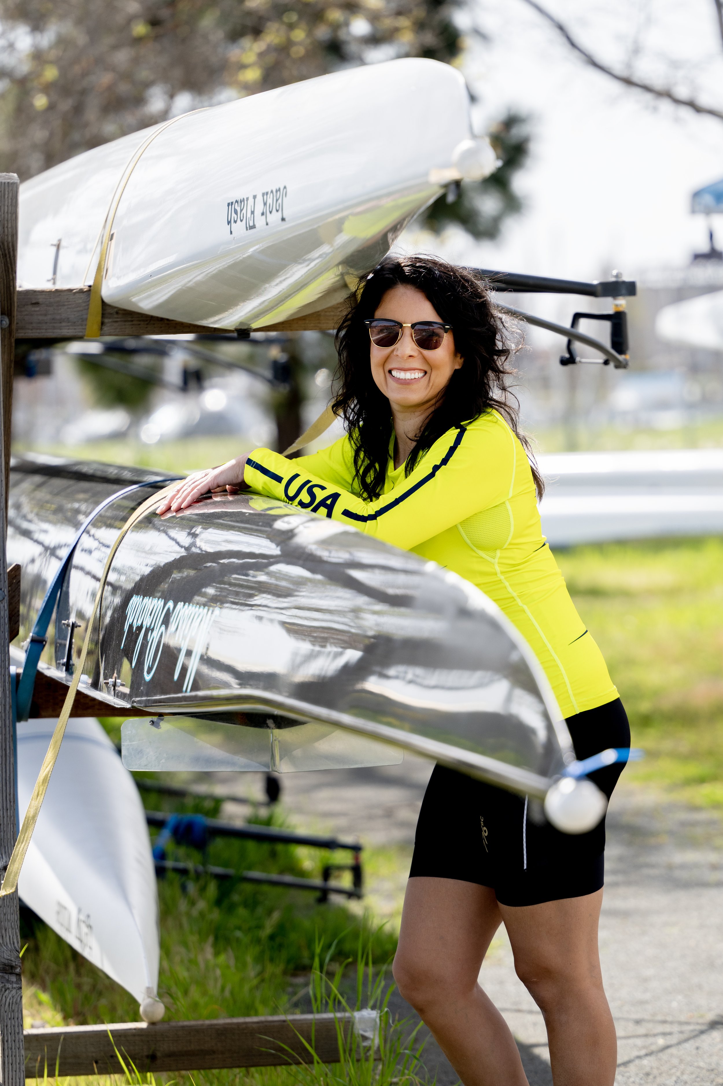 A woman wearing sunglasses and a yellow USA long-sleeve shirt smiling and leaning on a rowing shell boat that is mounted on a rack outdoors.