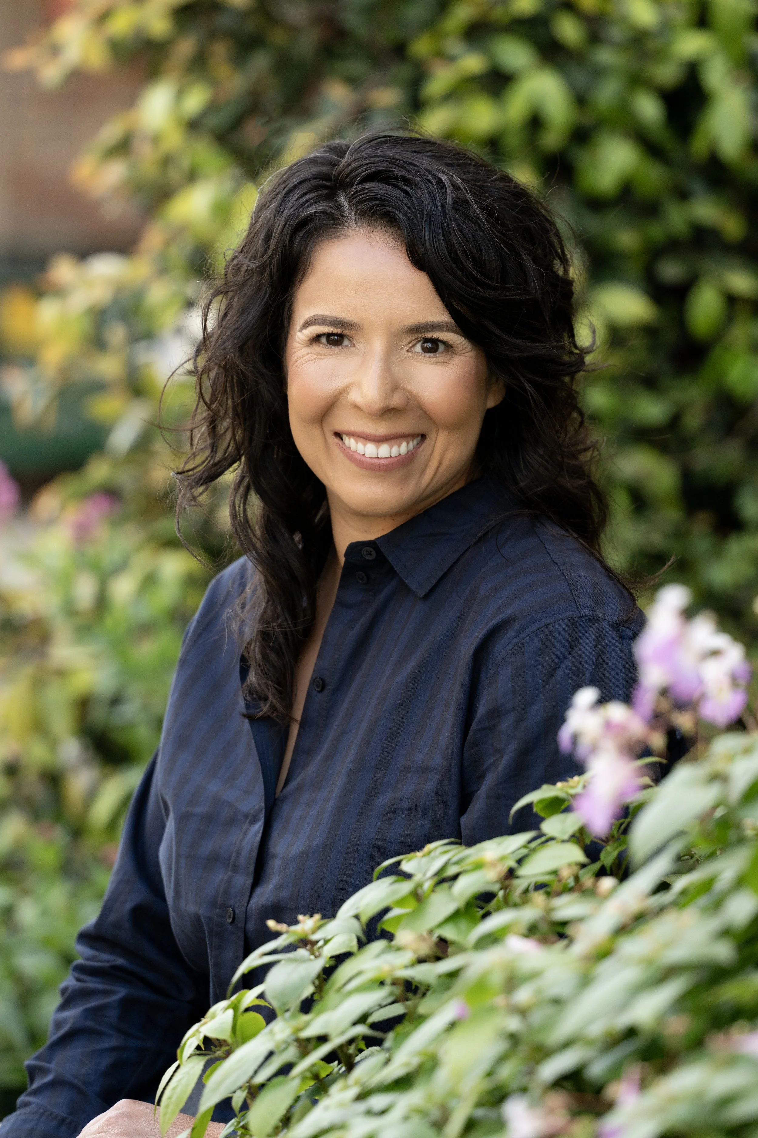 A woman with dark, wavy hair smiling outdoors among green and pink flowering bushes.