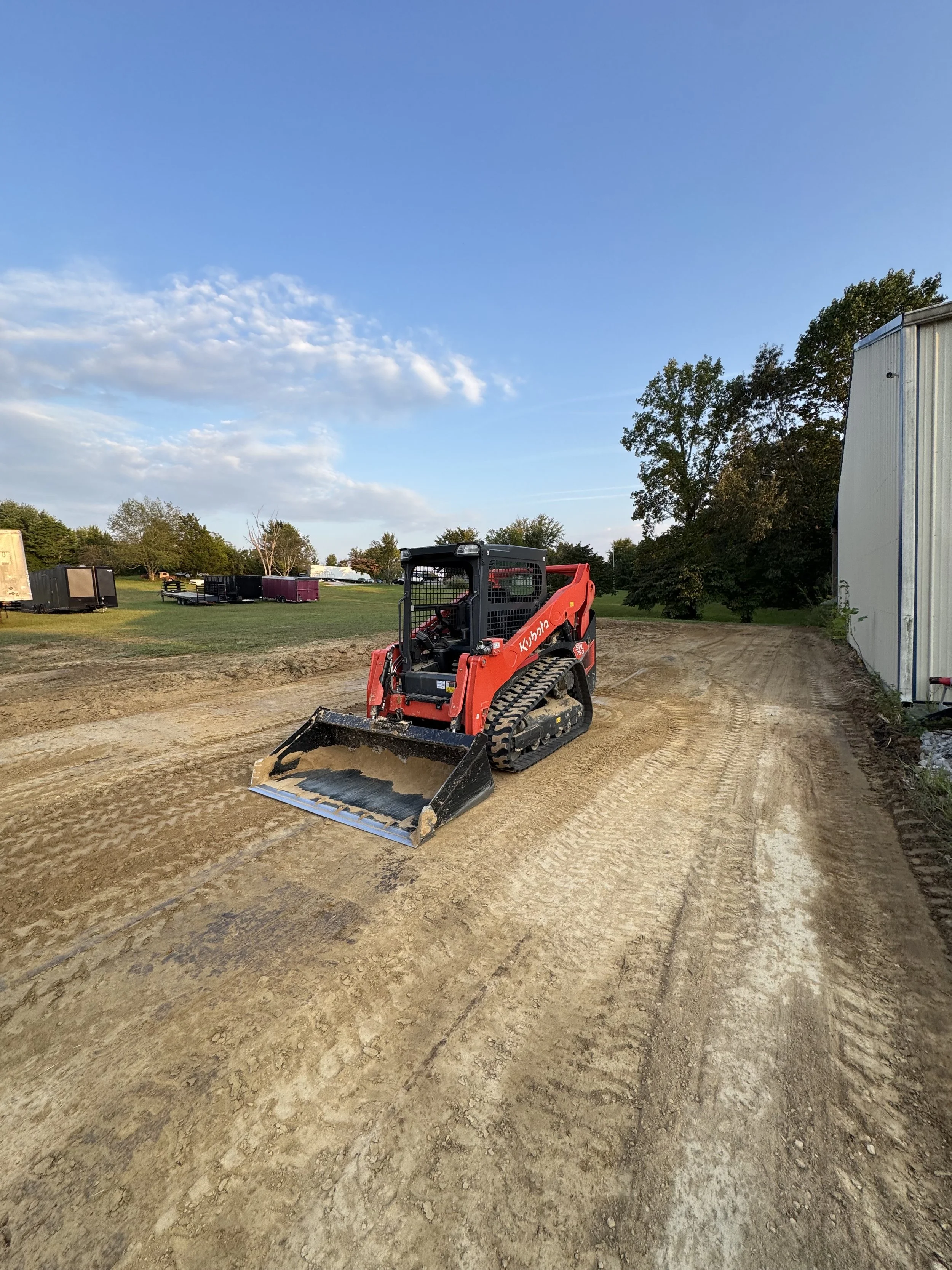 A red Kubota skid-steer loader on a dirt path at a construction site, with a clear blue sky and trees in the background.