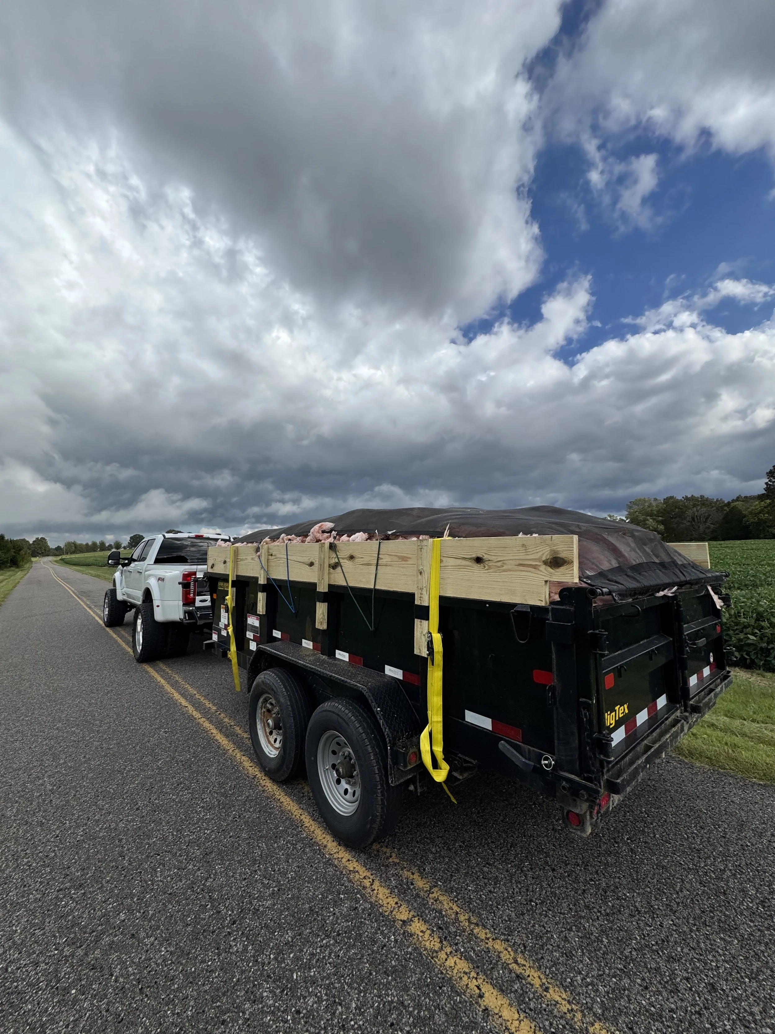 A white pickup truck towing a black trailer loaded with hay bales, parked on a rural road under a cloudy sky.