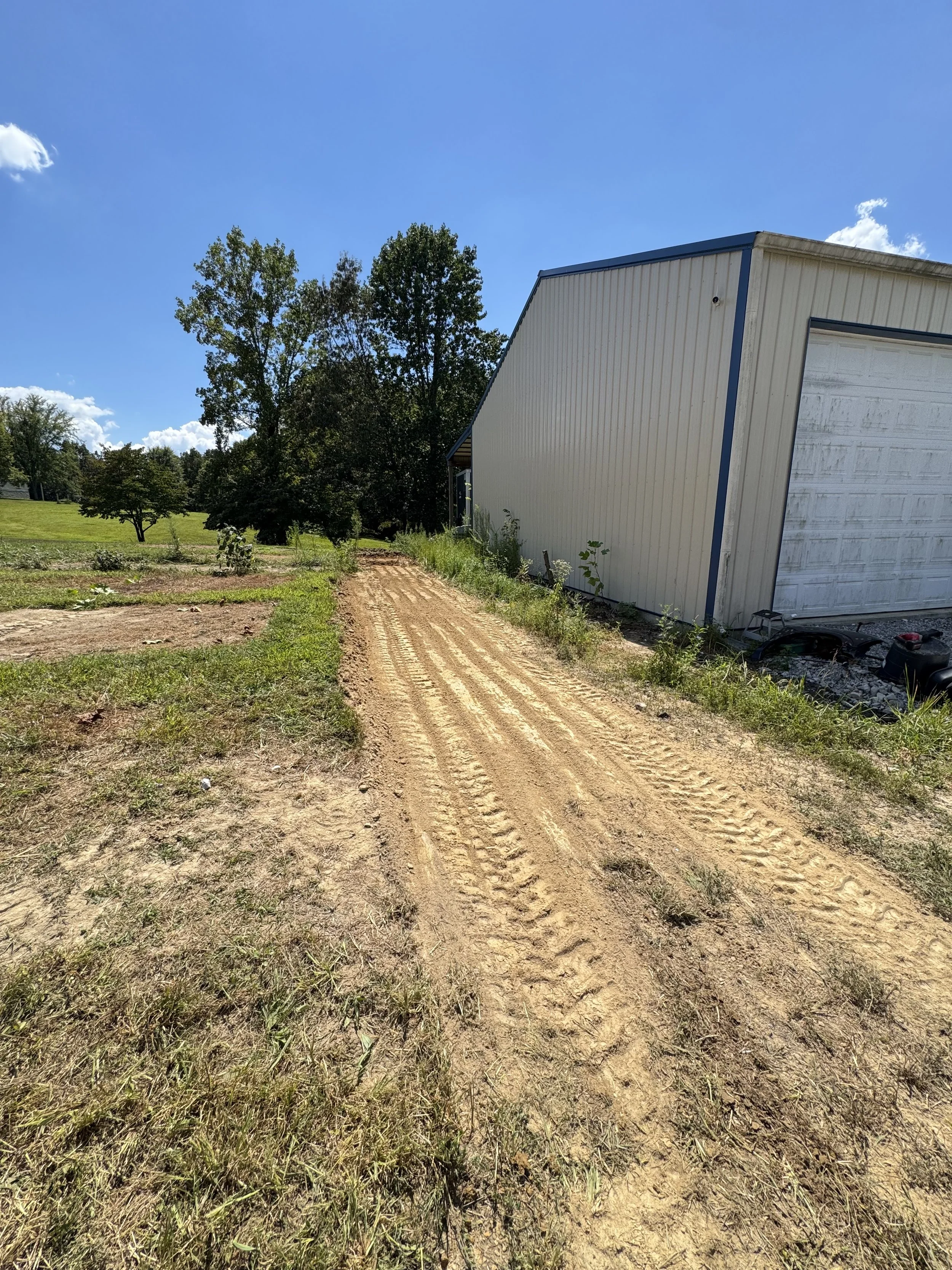 Dirt driveway next to a white building with a garage door, surrounded by grass and trees on a sunny day with a blue sky.