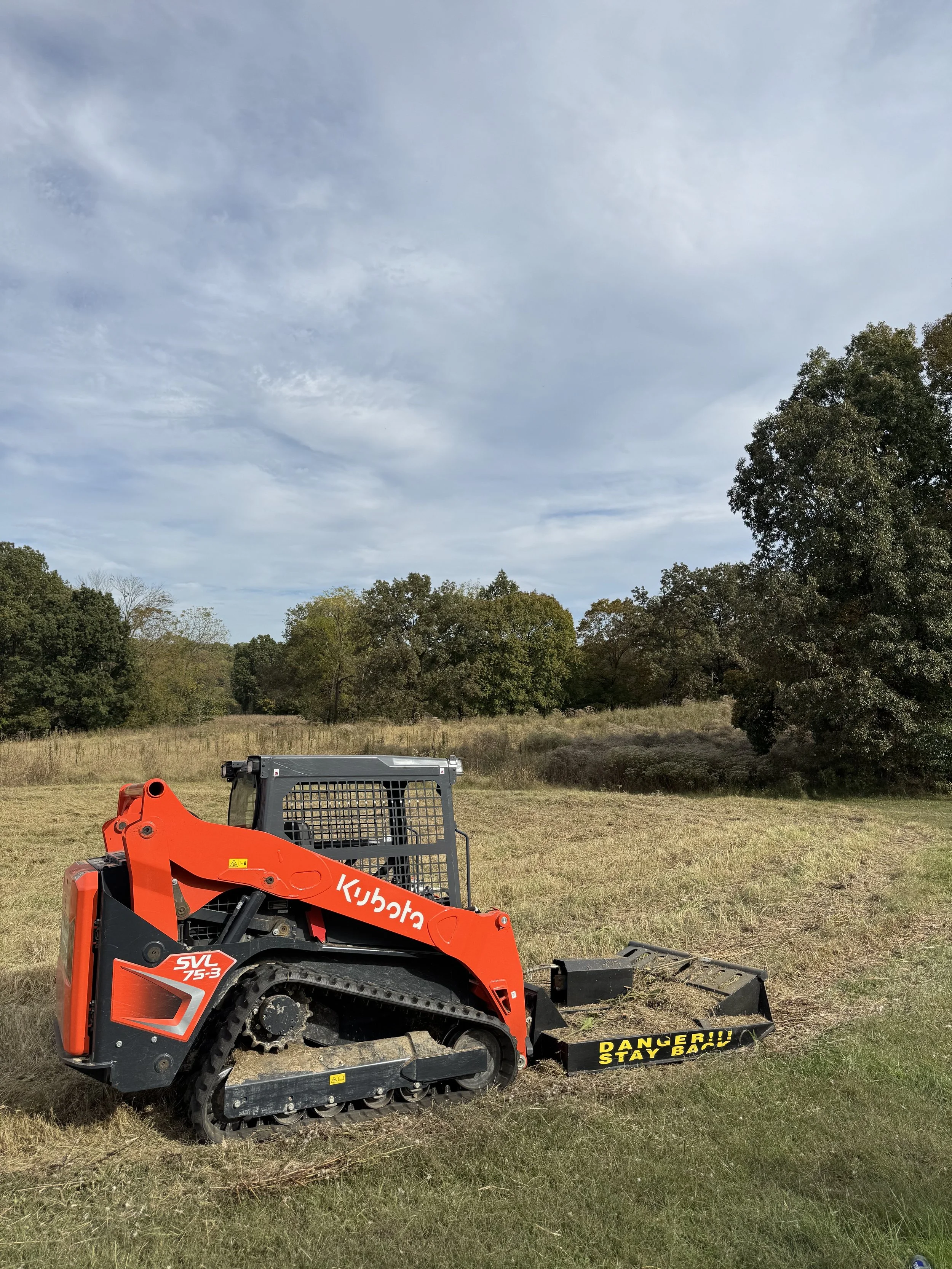 Orange Kubota compact track loader with a warning sign reading 'DANGER!! STAY BACK' on the attachment, parked on grassy field with trees and cloudy sky in the background.