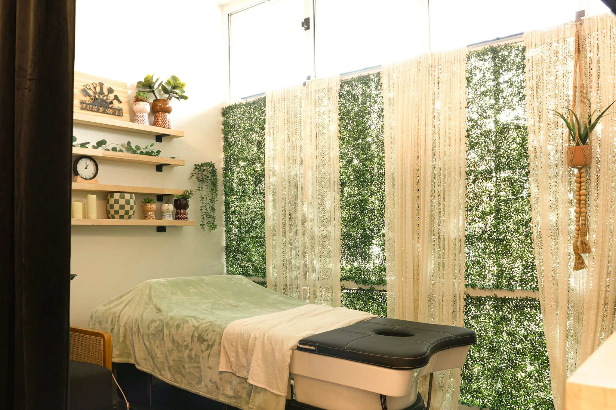 Massage therapy room with bed, green and beige decor, wooden shelves with plants and candles, lace curtains, and a treatment table.