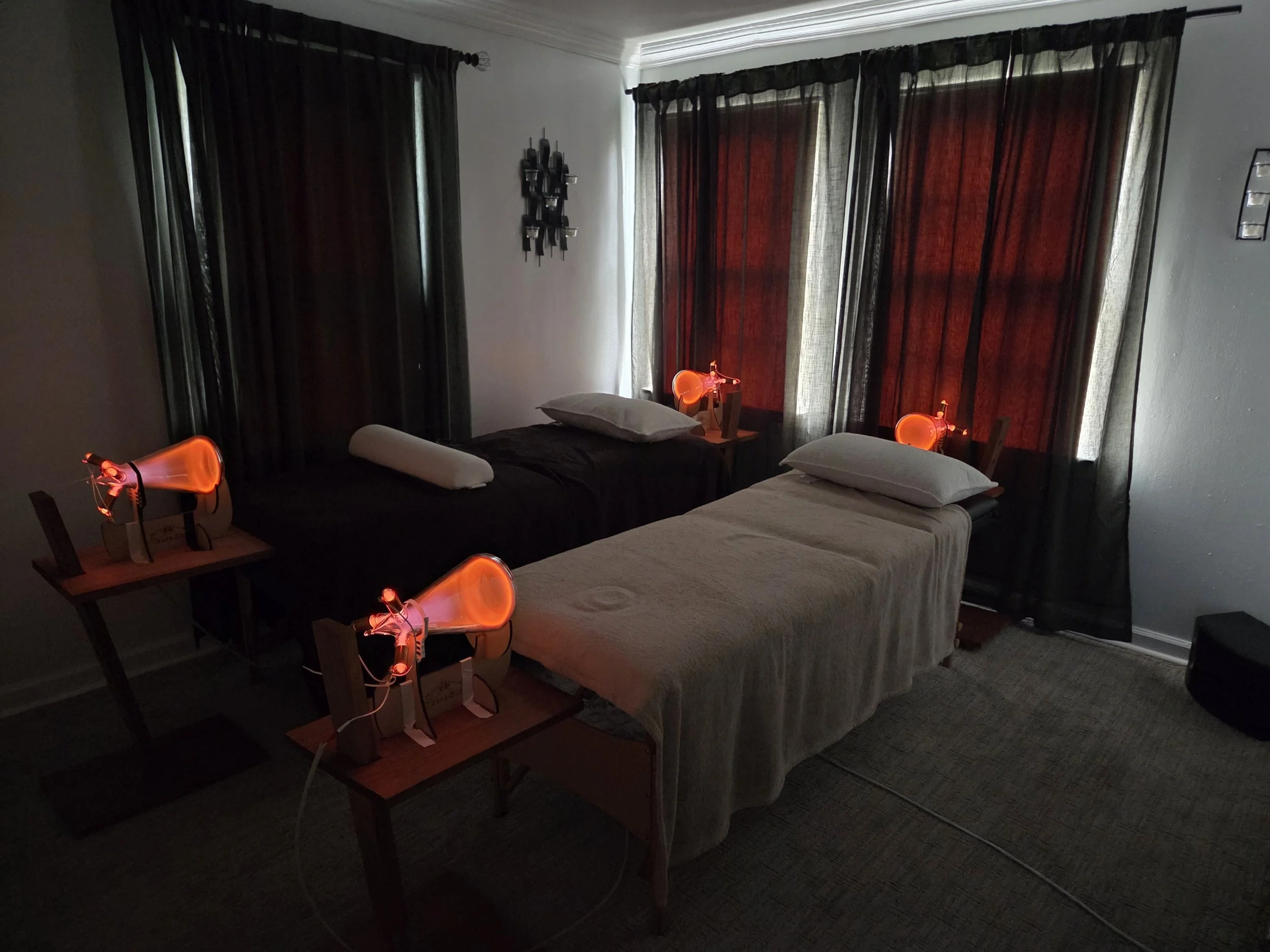 Room with two beds, each with a pillow and a towel, black and red curtains, and four orange heat lamps on small tables.