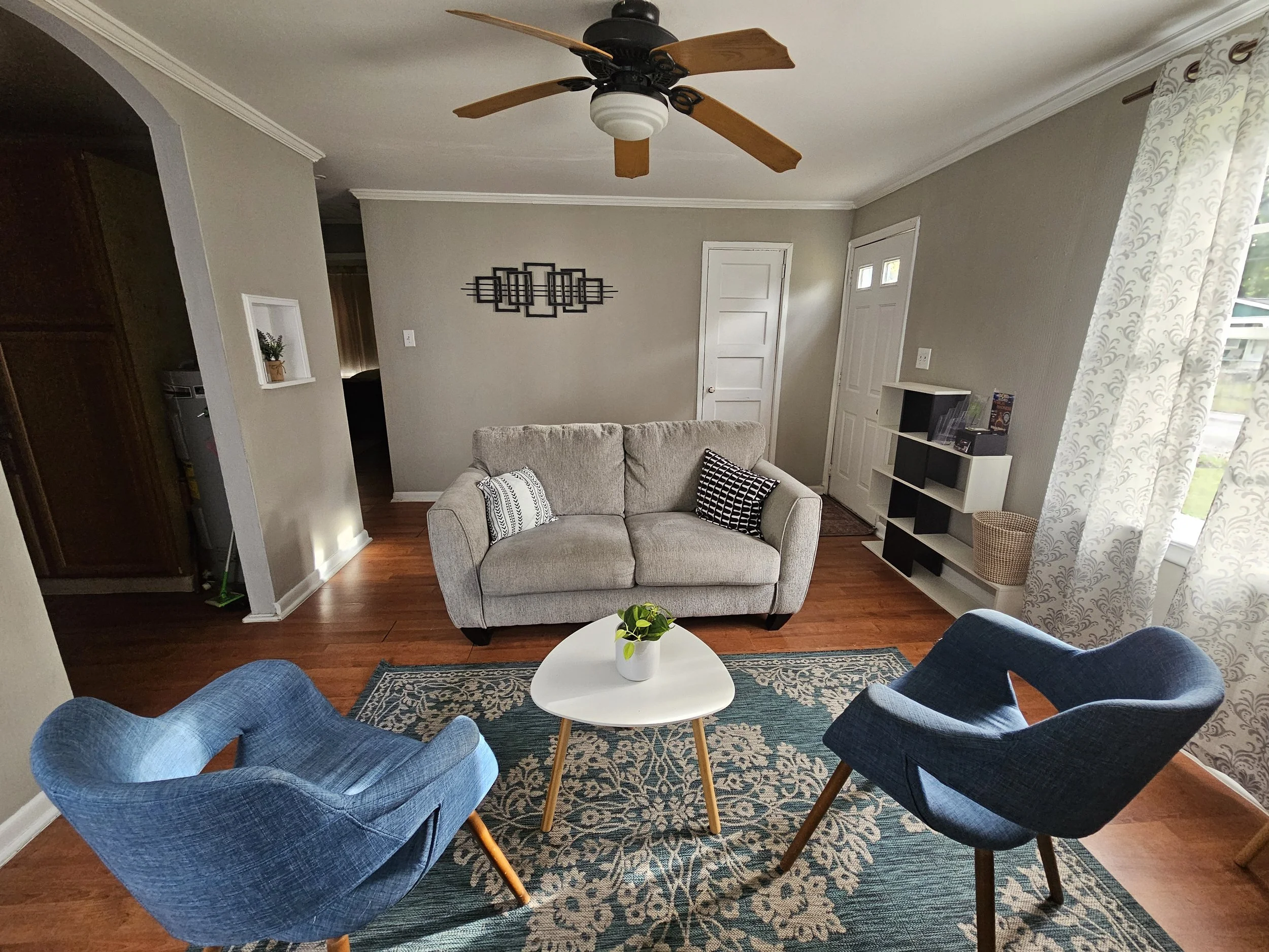 Living room with a light gray sofa, two blue armchairs, a white coffee table with a small plant, a patterned area rug, and a window with lace curtains. There's a ceiling fan and a wall-mounted shelf with books and a basket.
