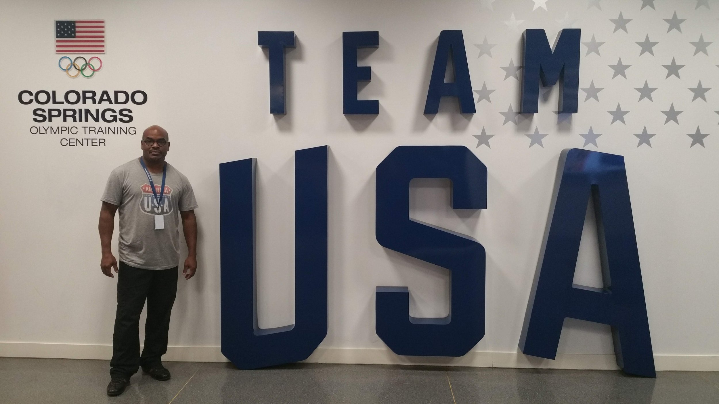 Man standing in front of large 3D letters spelling 'TEAM USA' at the Colorado Springs Olympic Training Center. The background features a wall with a pattern of stars, an Olympic logo, and the American flag.