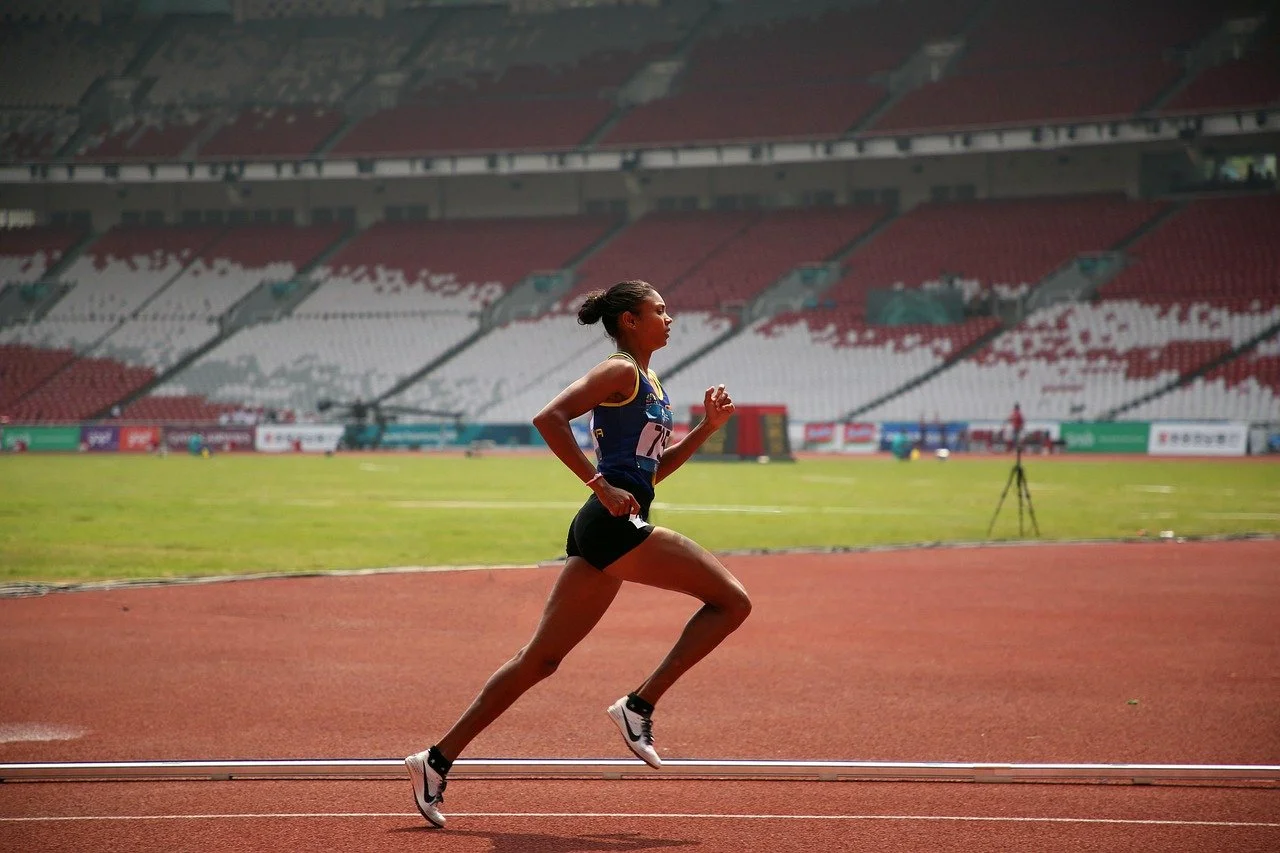 A female athlete running on a track in a stadium, wearing a blue top and black shorts.