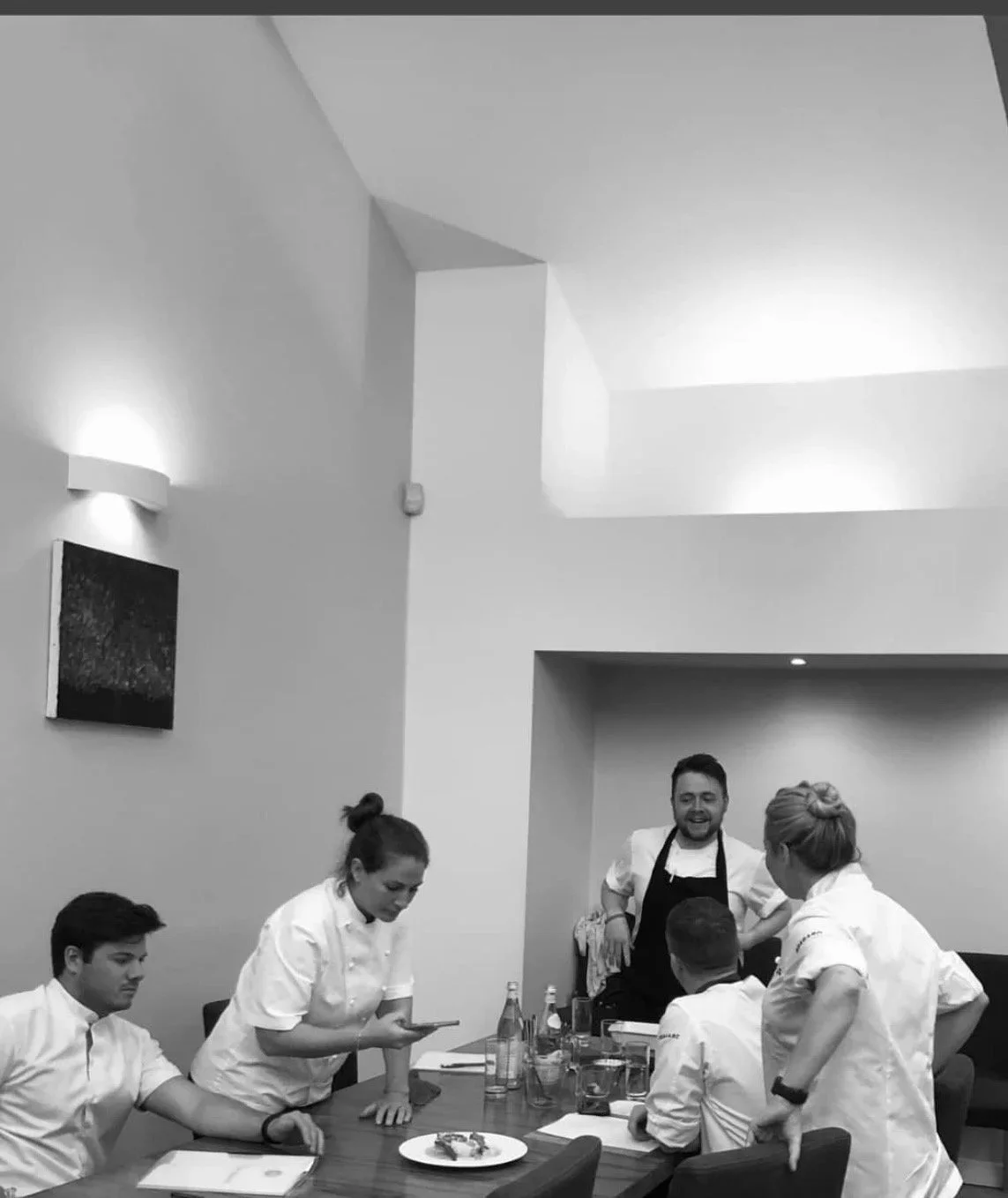 Group of five chefs in a kitchen, three women and two men, gathered around a dining table with food and drinks, engaged in conversation, with smiling expressions.