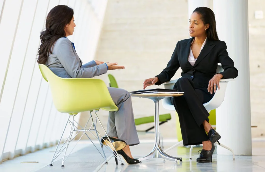 Two women having a conversation in an office, sitting across from each other at a small round table.