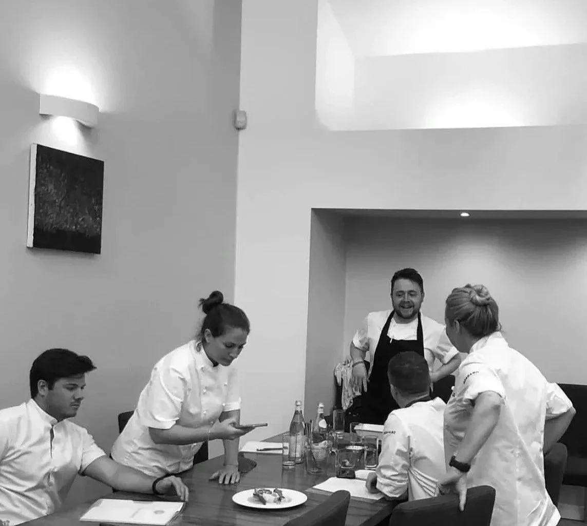 A group of five restaurant staff or chefs gathered around a table in a modern kitchen or dining area, engaging in conversation, with one person standing and smiling.