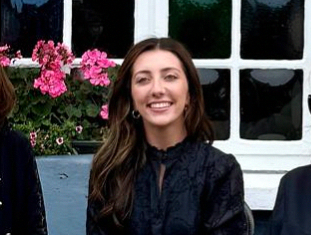A woman with long brown hair smiling at the camera, wearing earrings and a dark lace top, standing in front of a window with pink flowers outside.