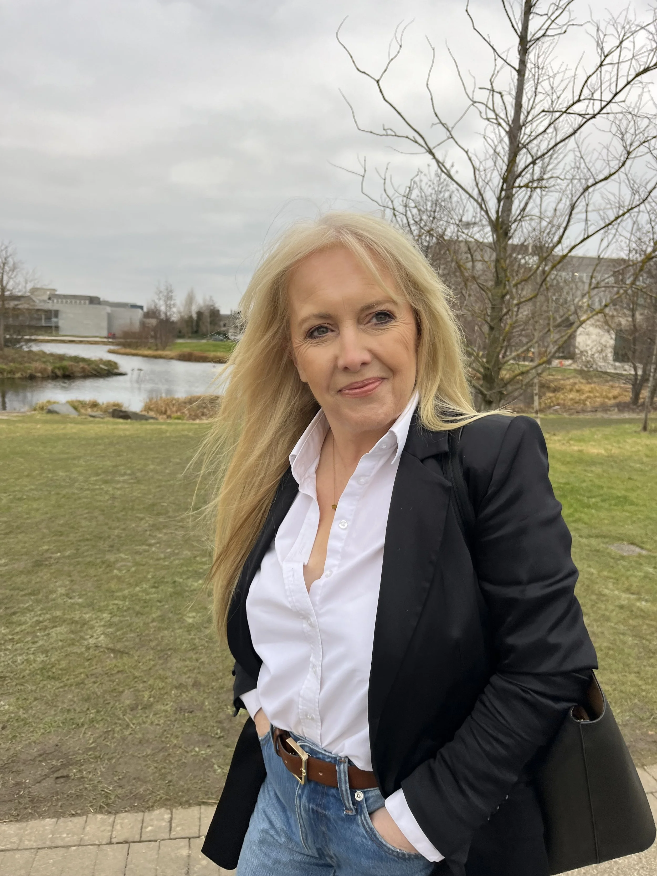 A woman with blonde hair wearing a white shirt and black blazer, standing outdoors by a pond with trees and buildings in the background.
