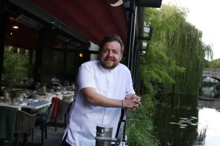 A smiling male chef standing in a kitchen with his hands on his hips, wearing a white chef's coat and a black apron, in black and white photograph.