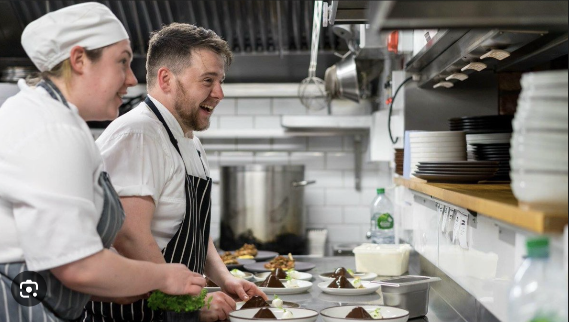 Two chefs, a woman and a man, smiling and preparing food in a commercial kitchen, with plates of plated dishes on the counter in front of them.