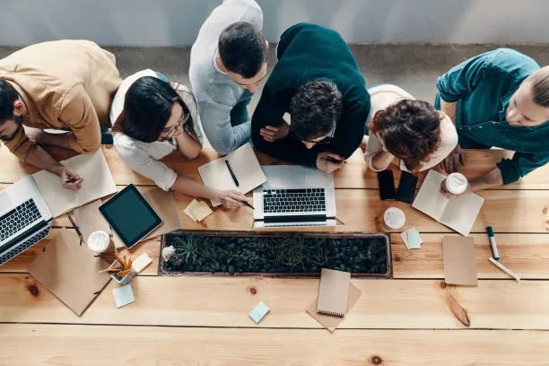 Group of six people gathered around a wooden table, looking at laptops and notebooks, engaged in a discussion or meeting.