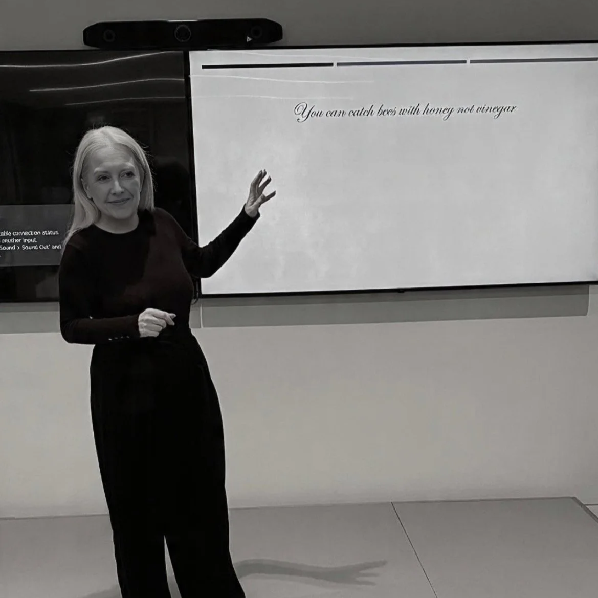 A woman in black clothing standing in front of a whiteboard with a quote: 'You can catch bees with honey not vinegar'.
