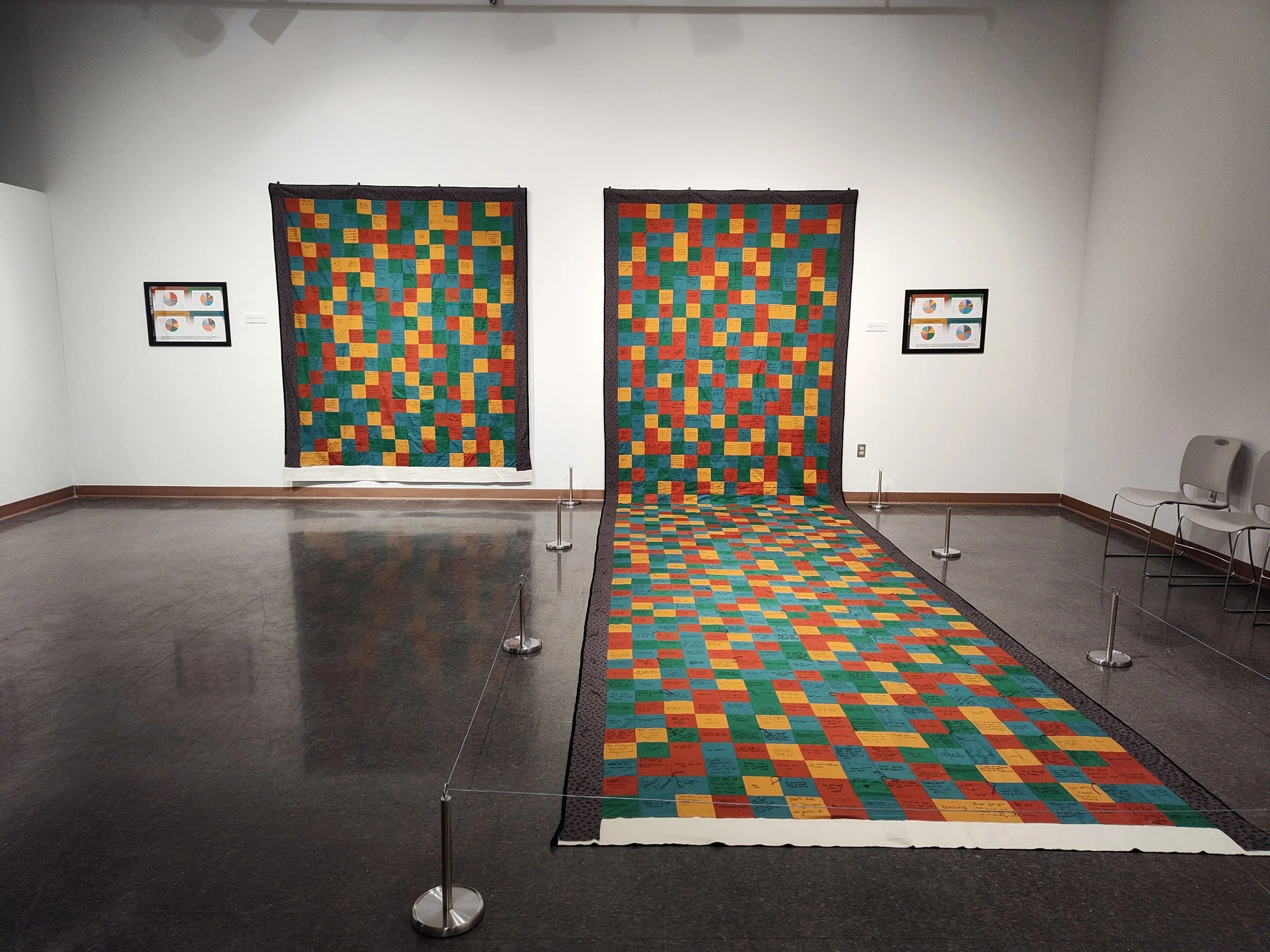 Colorful quilt art installation in a gallery, with two hanging quilts, with the one on the right draping over a large portion of the floor, surrounded by a barrier, with framed artwork on the walls.