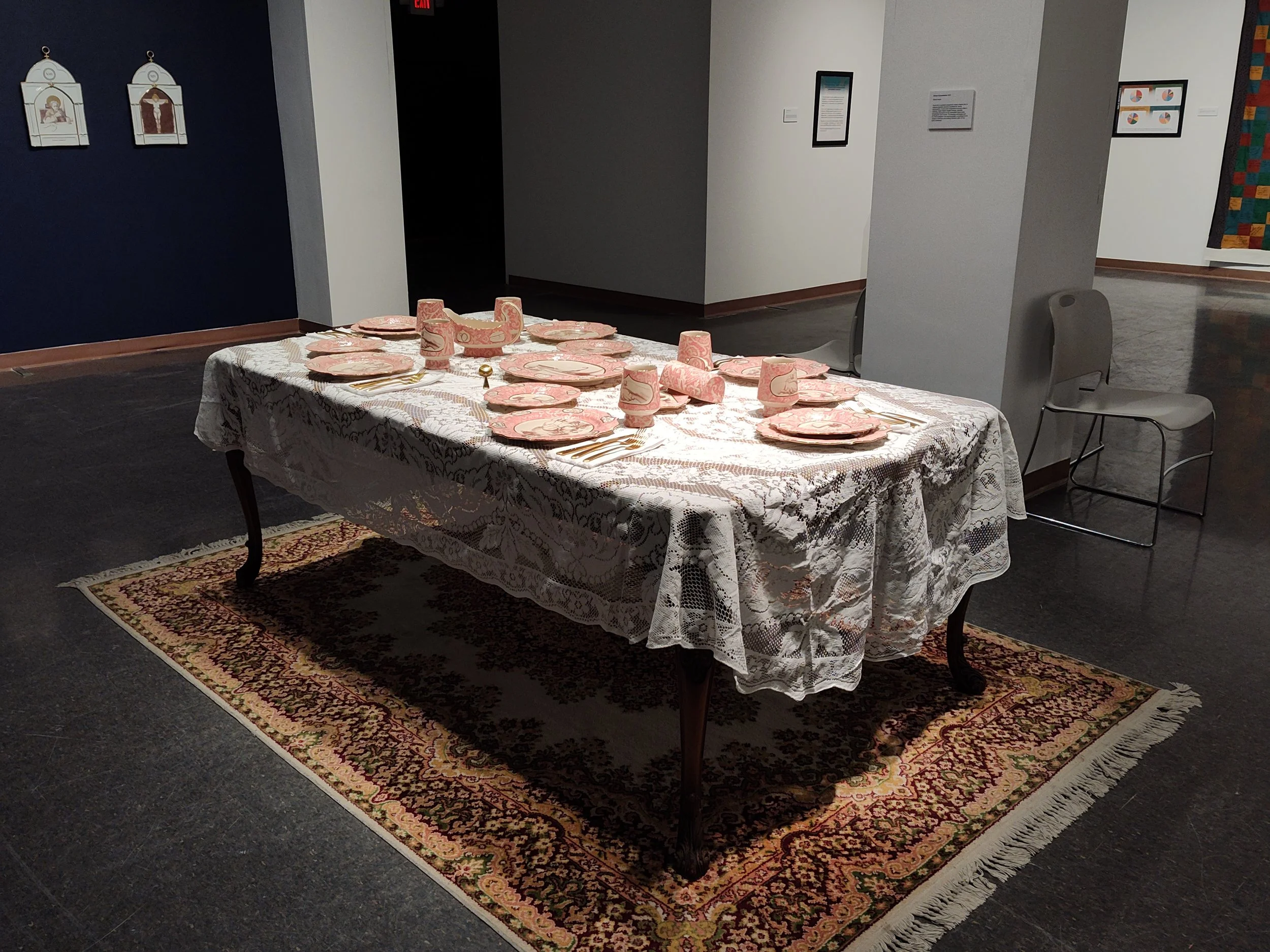 A dining table set with pink and white patterned plates, cups, napkins, and silverware, covered with a lace tablecloth, on a decorative rug in an art gallery.