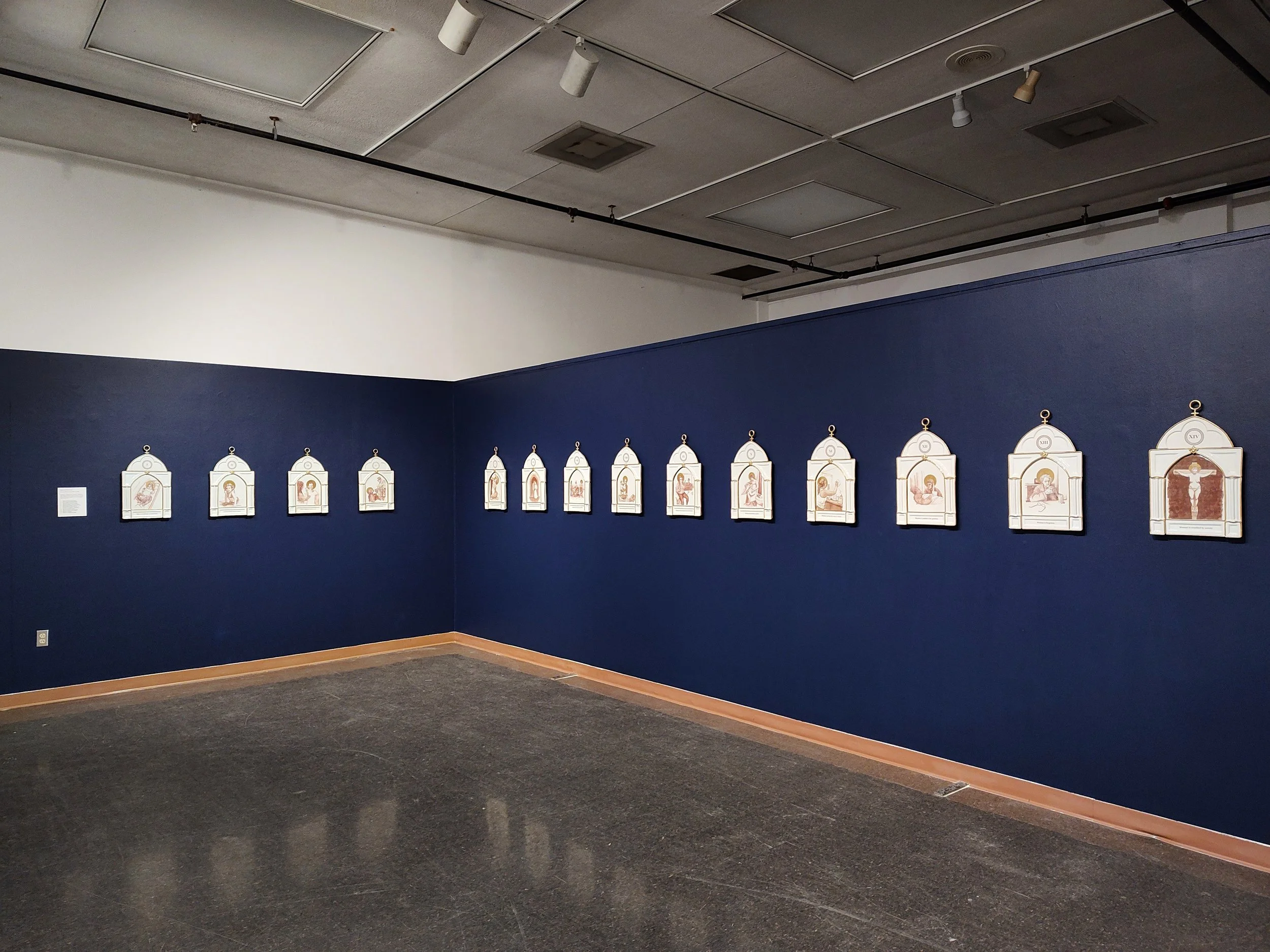 An art gallery with a dark blue wall displaying a series of religious or iconographic illustrations on white plaques. The ceiling has exposed panels and track lighting, and the floor appears to be dark concrete.