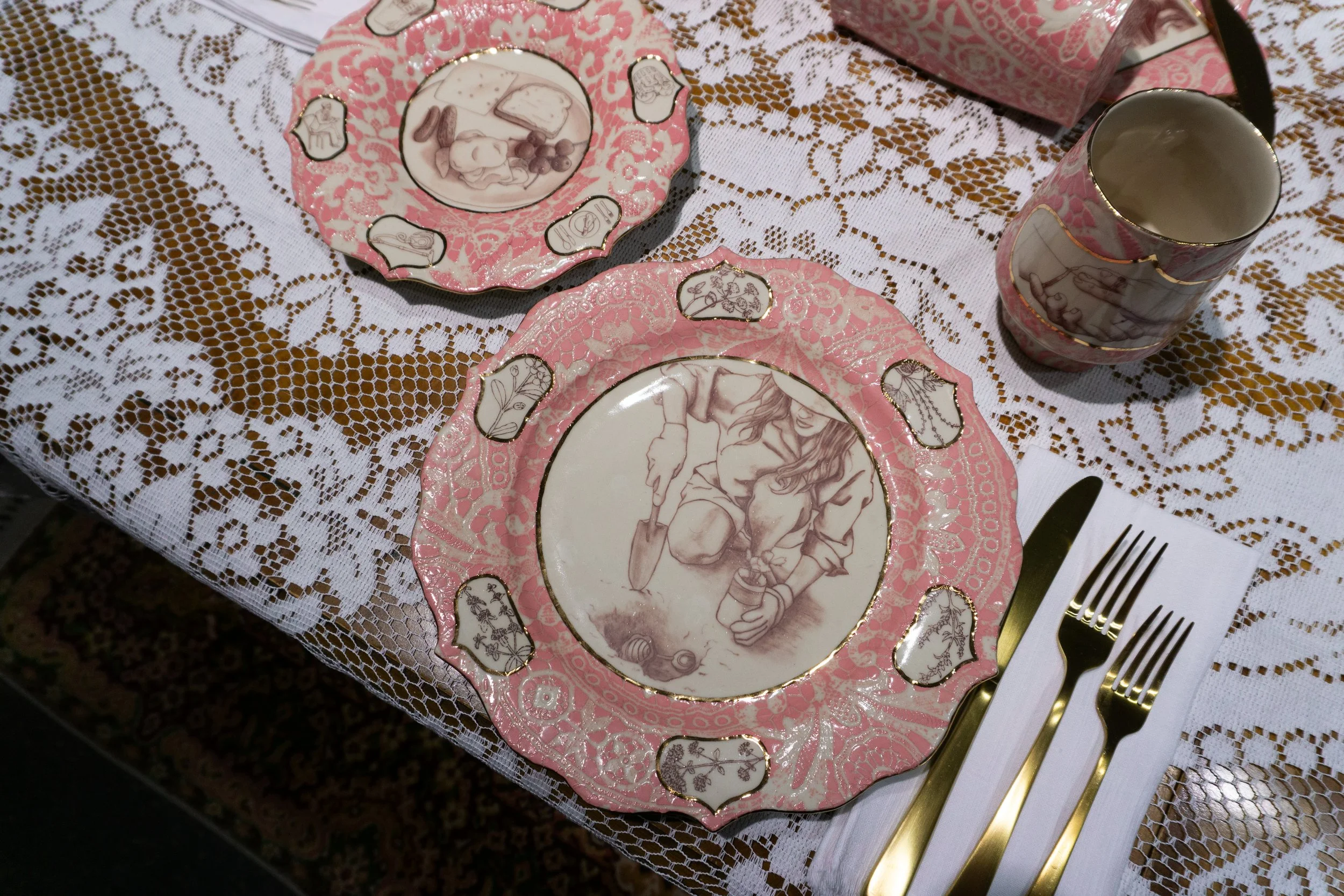 A table setting with decorative pink and gold china plates, a matching mug, and gold utensils on a white napkin. The plates have vintage illustrations and patterns, with one featuring a woman planting a seed.