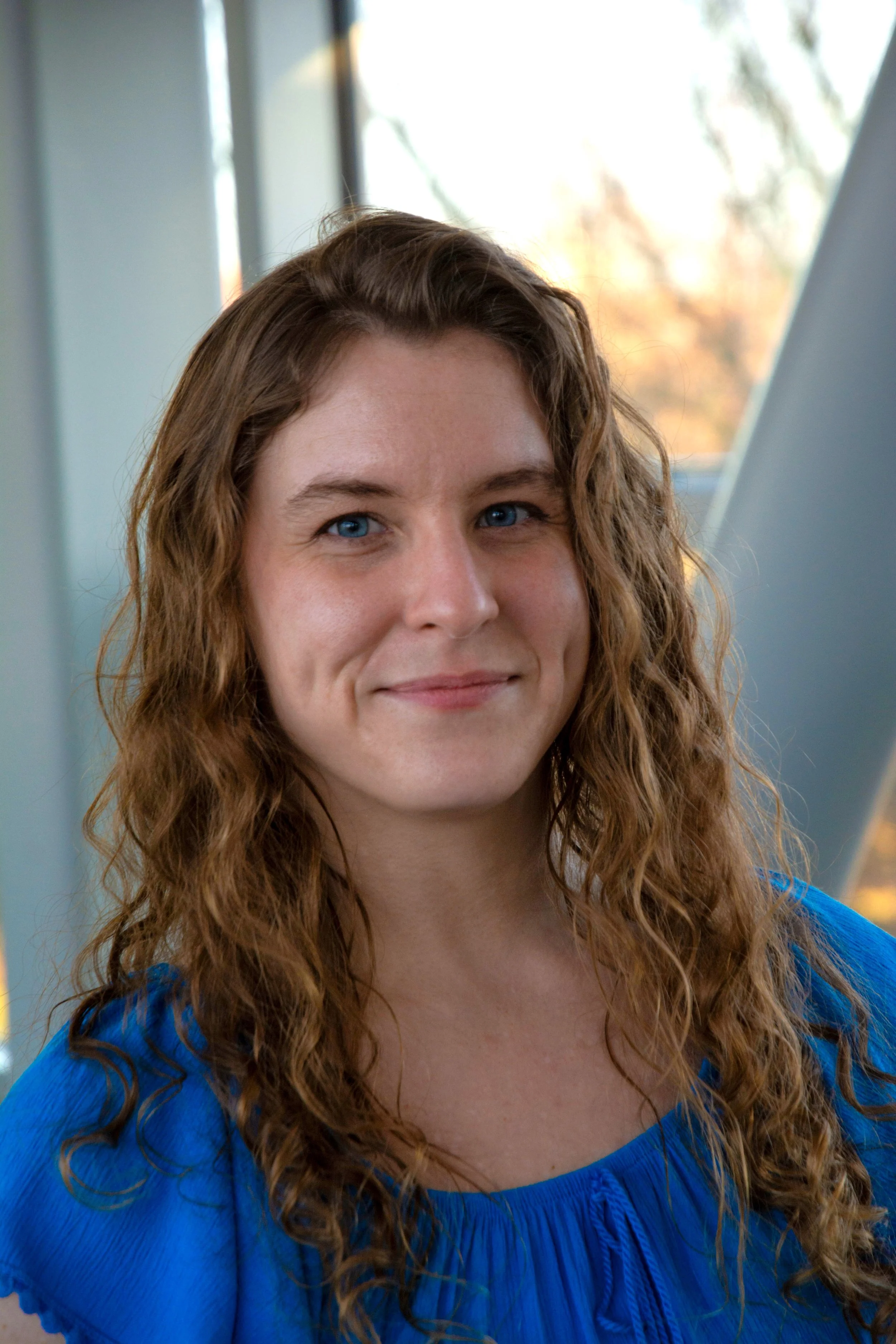 A woman with long, curly red hair and blue eyes in a blue top, smiling indoors near a window with a blurred outdoor background.