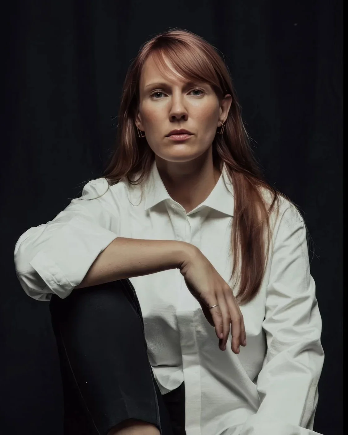 Portrait of strategist and educator, Baiba Matisone, with red hair in a white shirt sitting against a dark background.