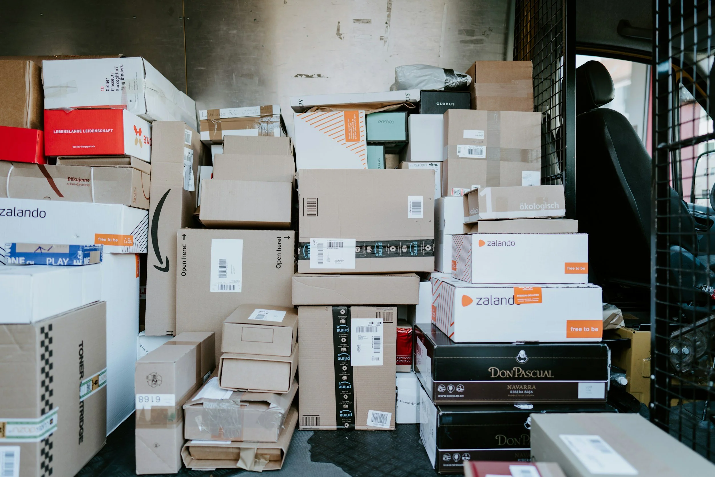 Stacked cardboard boxes in a warehouse or delivery truck with various logos and labels, including Amazon, Zalando, Don Pascual, and others.