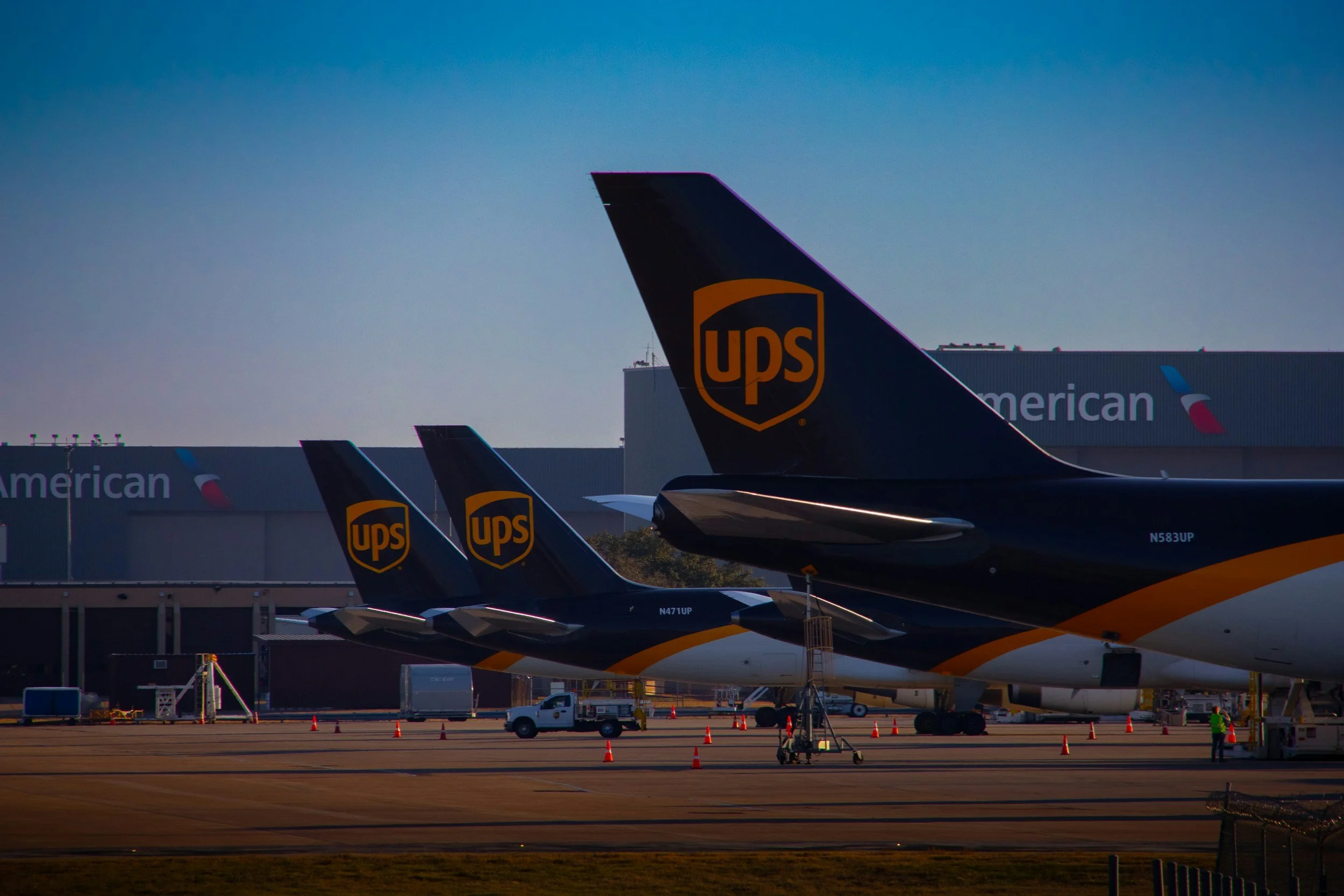 Three UPS cargo planes parked on the tarmac at an airport with American Airlines hangar in the background.