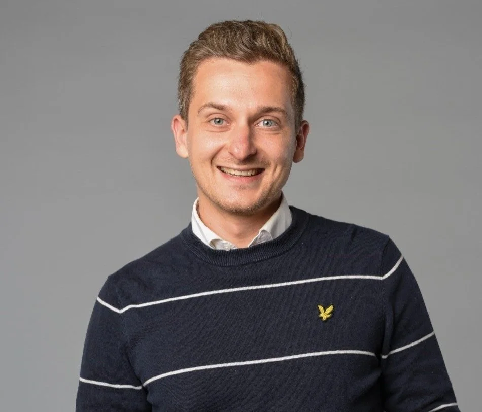 A young man with light brown hair and blue eyes, smiling, wearing a navy blue sweater with white horizontal stripes and a yellow emblem, over a collared shirt, against a gray background.