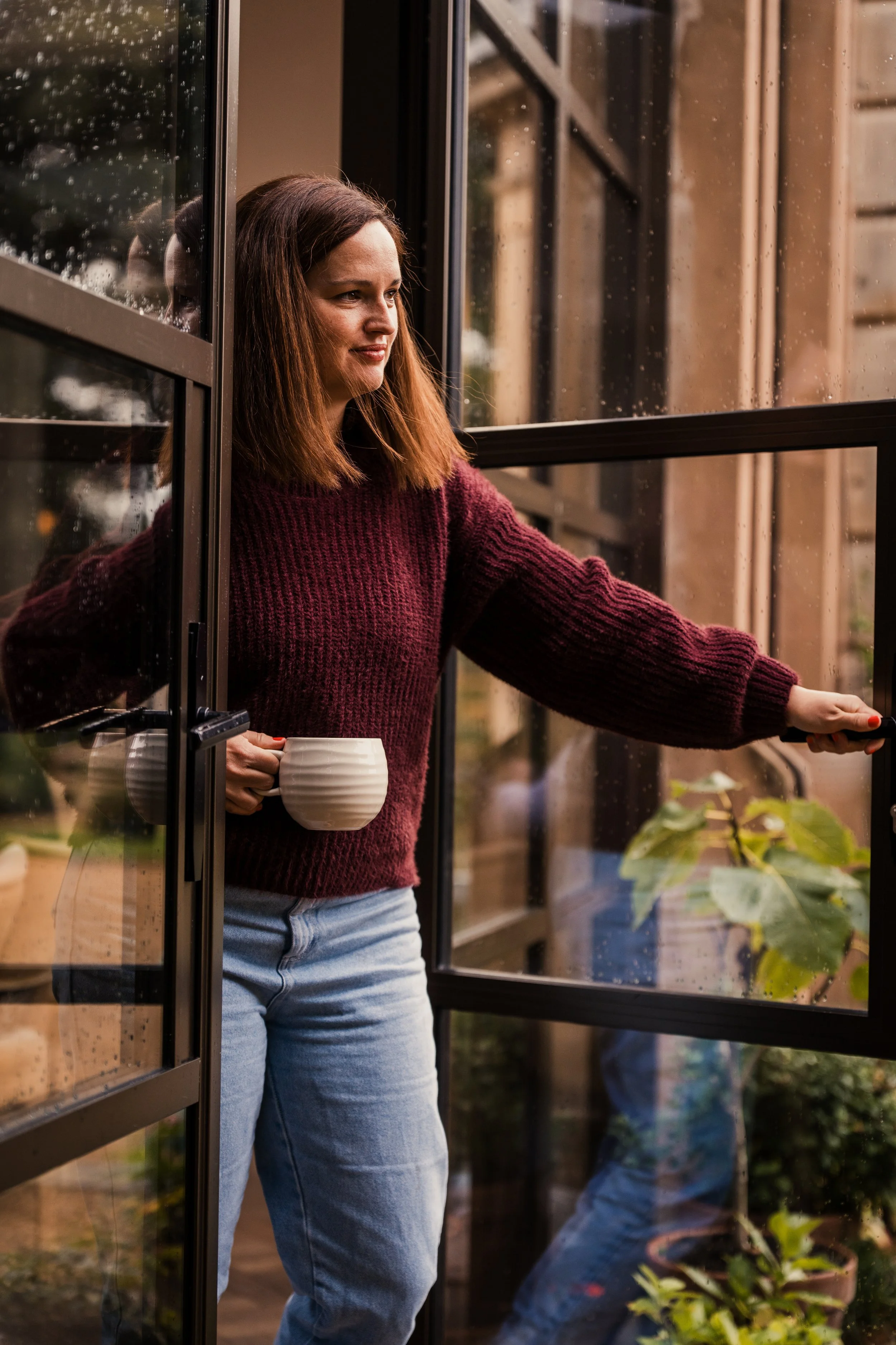 A woman with shoulder-length brown hair, wearing a maroon sweater and light blue jeans, stands at an open glass door holding a white mug in her right hand. She is looking outside, surrounded by a glass structure with visible rain droplets and potted plants nearby.