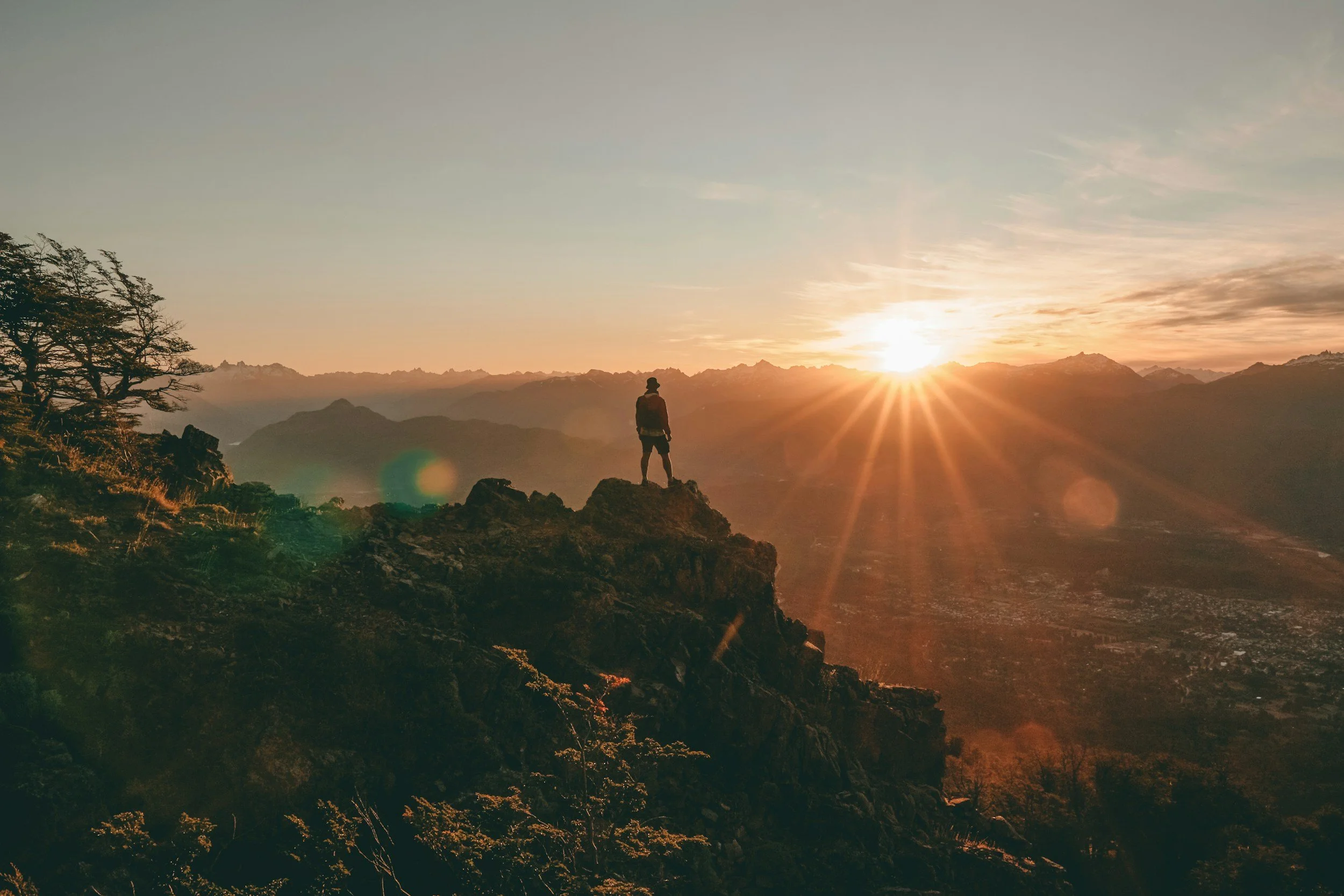 Person standing on a rocky mountain ridge during sunset with a mountain range in the background.