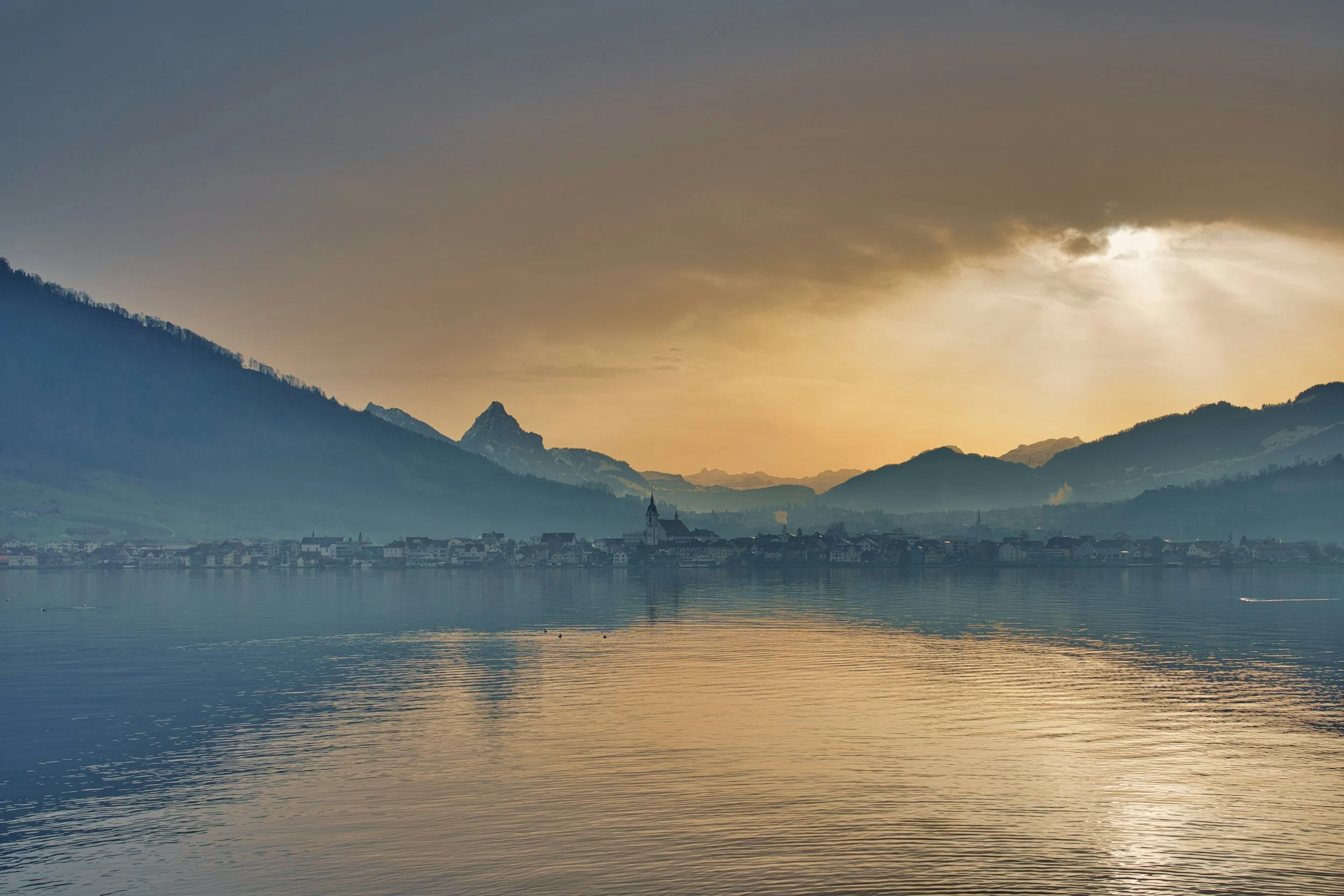 A serene lake with gentle ripples reflecting the sunset sky. In the background, a town is nestled along the shoreline, surrounded by mountains, some with snow-capped peaks. The sky is partly cloudy with sunlight breaking through.