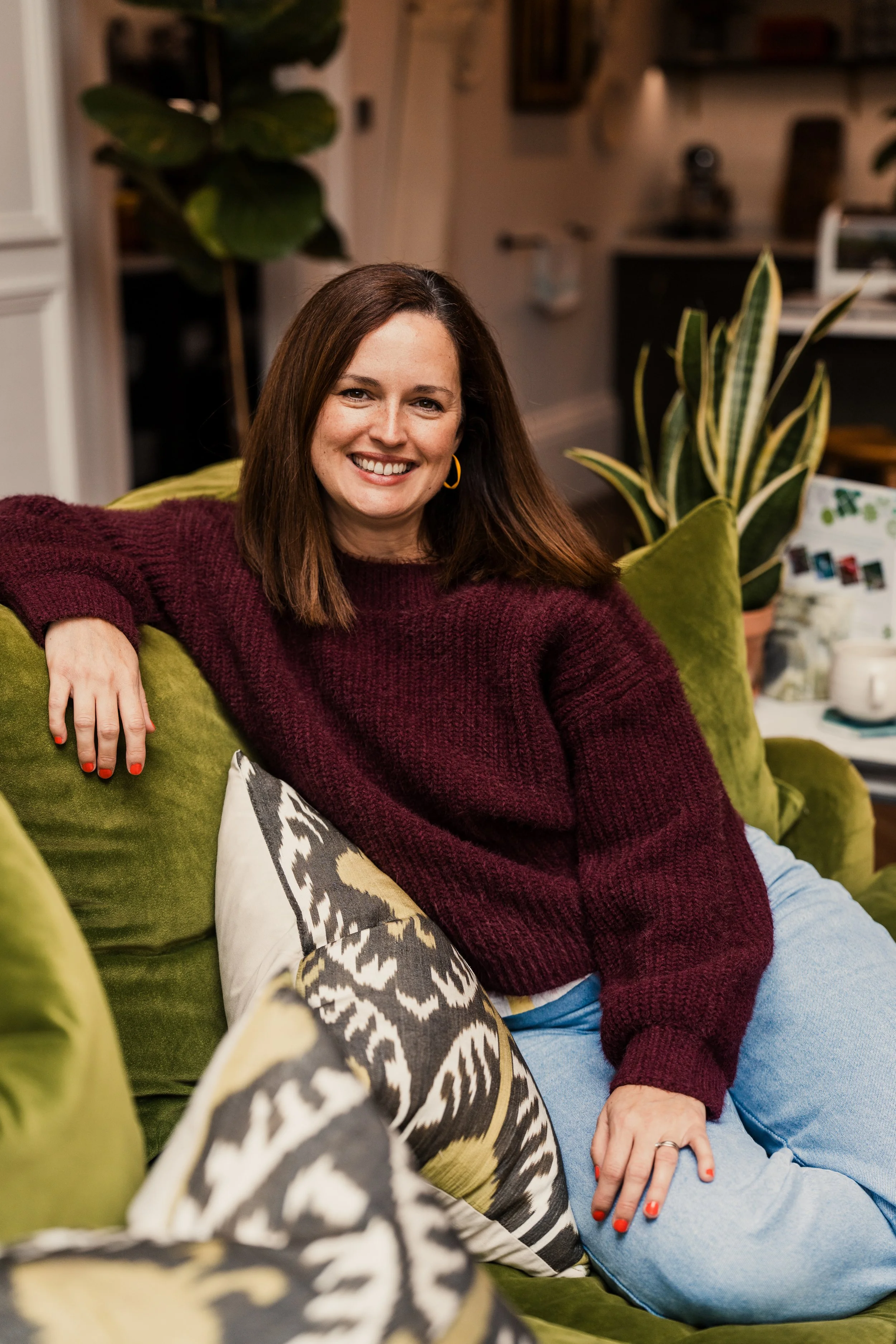 A woman with shoulder-length brown hair, wearing a burgundy sweater, light blue jeans, and gold hoop earrings, sitting on a green sofa in a cozy living room. She is smiling and leaning back with her right arm resting on the sofa's backrest, surrounded by patterned pillows and potted plants, with a blurred background of kitchen and wall decorations.